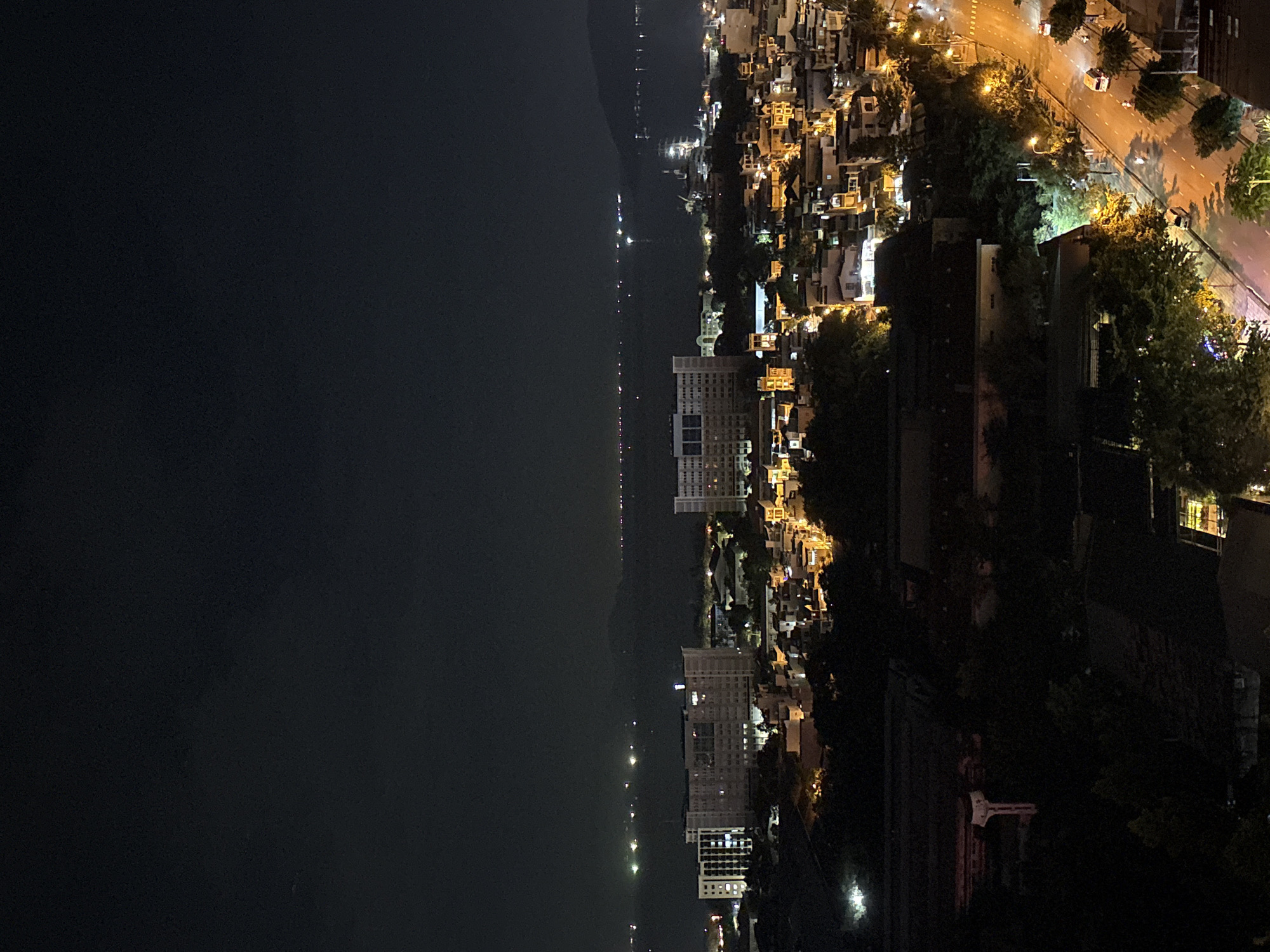 The image depicts a night-time aerial view of a city, likely Nha Trang, Vietnam, with a focus on urban development extending towards a body of water. The foreground is dominated by dark foliage and some residential structures, with a prominent road cutting through the lower right quadrant of the image. This road is illuminated by streetlights and shows a few vehicles, including cars and motorcycles, in motion. The middle ground reveals a densely packed urban area with numerous buildings of varying sizes, many of which are lit up, indicating activity. Prominent among these buildings are several large, multi-story structures, including what appears to be a hospital or institutional building with a distinct illuminated sign at its top. Further in the distance, the city lights blend with the reflections on the water, suggesting a coastal or bayside location. The horizon is marked by the dark silhouette of hills or mountains in the far distance, with faint lights scattered across the water, possibly from boats or distant coastal settlements. The overall atmosphere is that of a vibrant, active city at night.