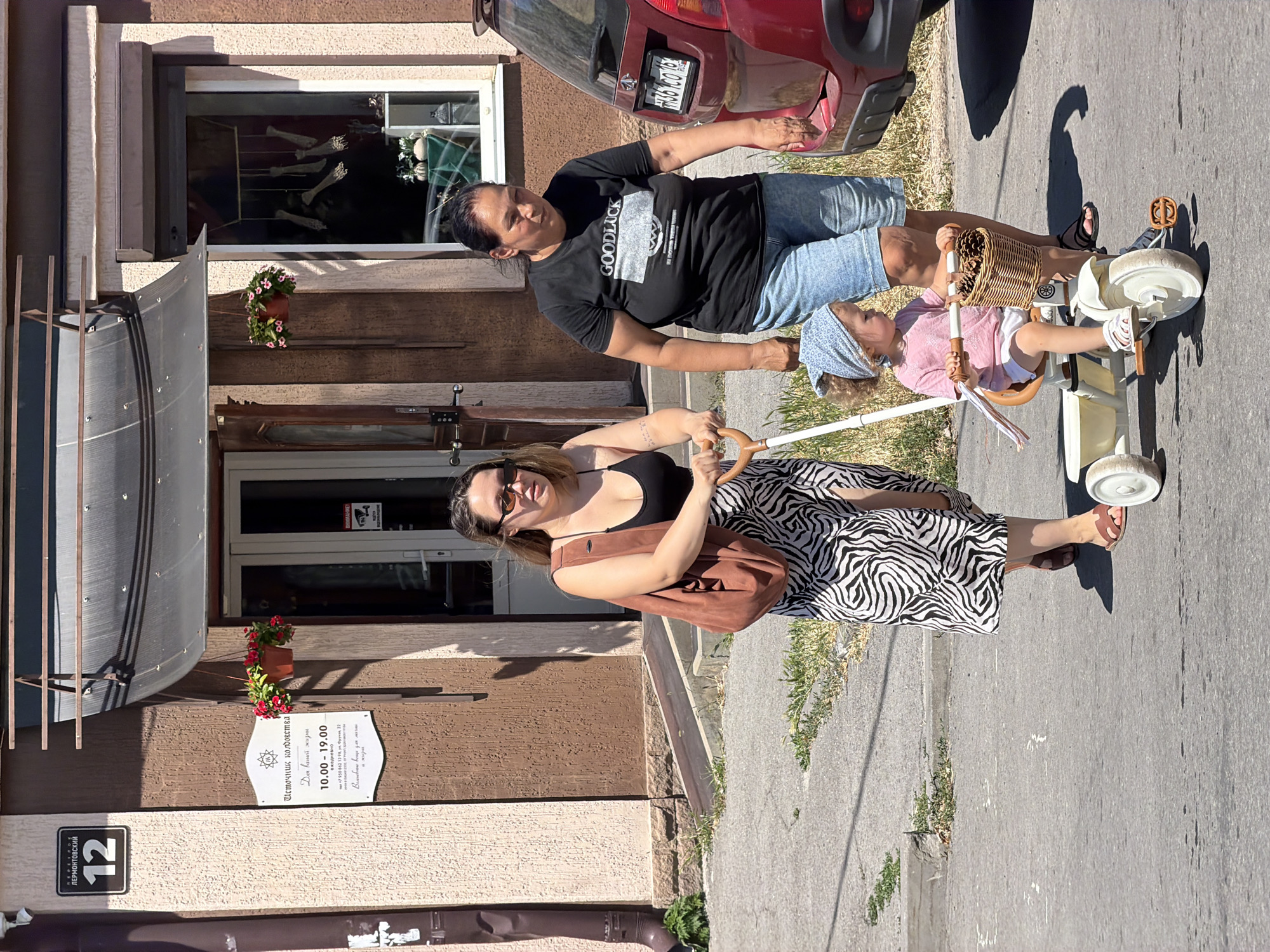 Two women and a young child are walking on a sidewalk in Taganrog, Russia, on a sunny day. The woman on the left is wearing a black tank top, sunglasses, a long zebra-print skirt, and brown sandals. She is carrying a large brown bag over her shoulder and pushing a tricycle with the child. The child, wearing a pink shirt, white shorts, and a blue bandana, is sitting on the tricycle, which has a wicker basket on the front. The woman on the right is wearing a black t-shirt with "GOODLUCK" printed on it, denim shorts, and black sandals. She is walking next to the tricycle.
Behind them is a building with a brown facade and a dark awning over the entrance. A sign with Russian text and a building number "12" is visible on the wall. There are flower pots with red flowers hanging near the entrance. A red car is parked on the right side of the image. The ground is paved with asphalt and concrete.