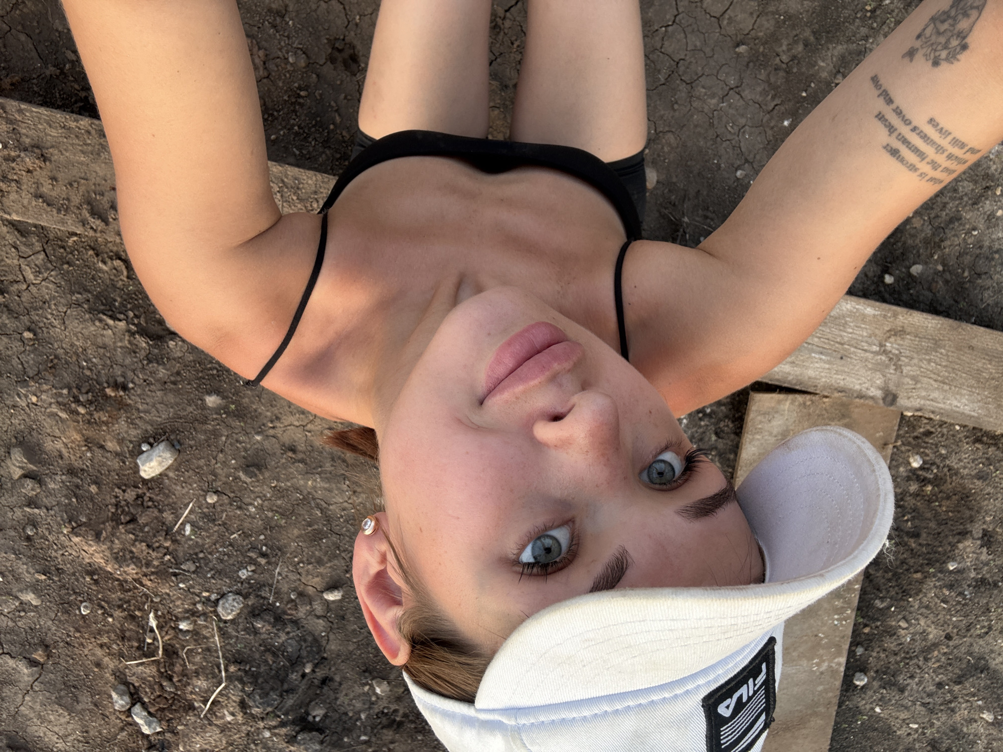 This is a medium shot, overhead, selfie-style photograph of a young woman lying on cracked, dry, dirt ground. She is wearing a black spaghetti strap top and a white baseball cap with a Fila logo. Her arms are extended upwards and out to the sides, with a tattoo visible on her left arm. Her face is tilted towards the camera, with bright blue eyes and light pink lipstick. She has a small diamond stud earring in her visible right ear. The scene suggests an outdoor, possibly rural setting during the daytime, with harsh sunlight indicated by the shadows and the cracked earth. The image appears to be taken in Лобаново, Russia. The text on her arm is a quote that reads, "What is stronger than the human heart which shatters over and over and still lives."