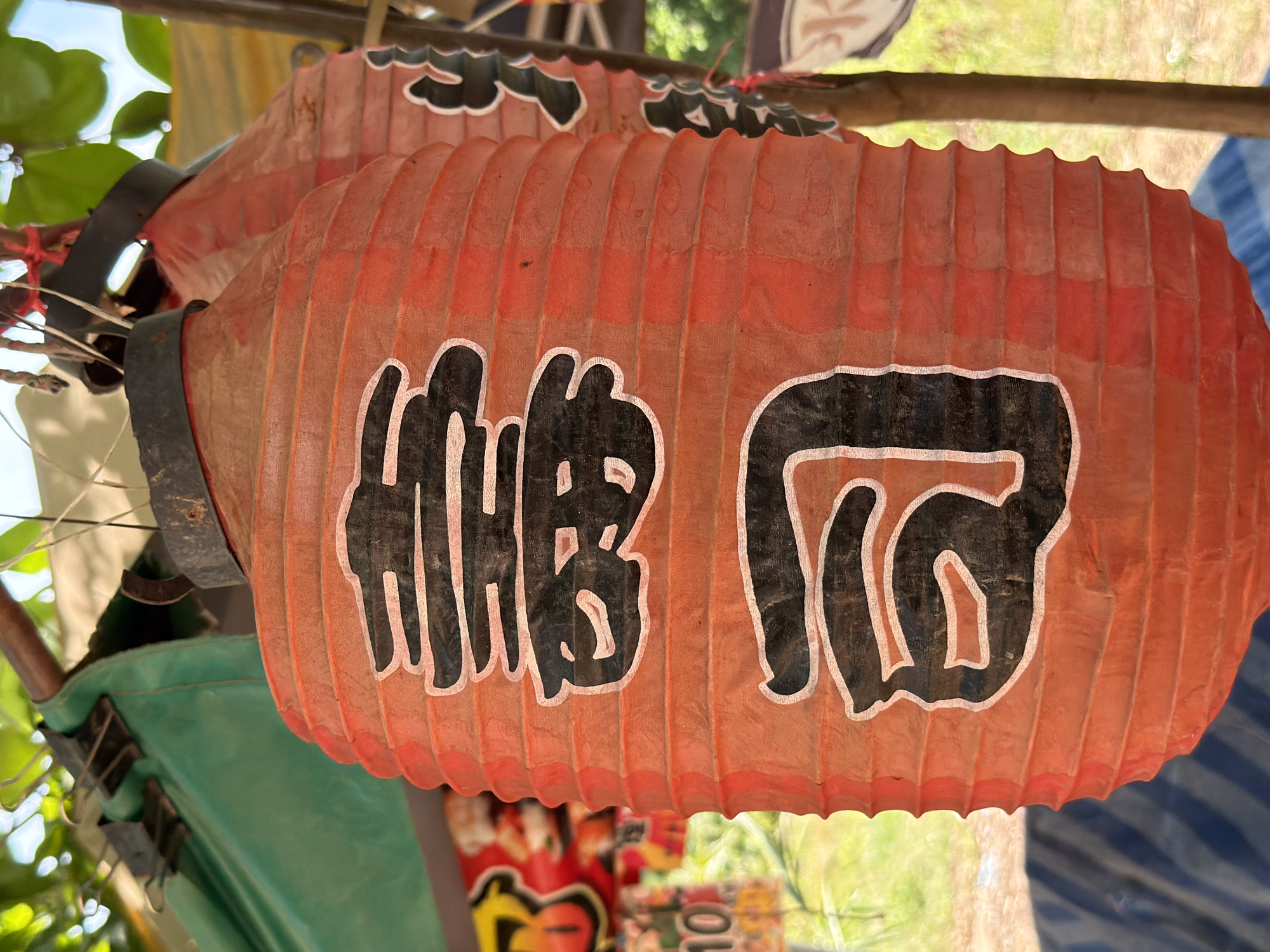 The image shows two red Japanese paper lanterns hanging outdoors in what appears to be a market stall in Baan Bo Phut, Thailand. The lantern in the foreground has black Japanese characters painted on it. The lantern is cylindrical with horizontal pleats. The top of the lantern has a black band. In the background, there is a green tarp, some other merchandise, and foliage. The lighting suggests it is daytime.