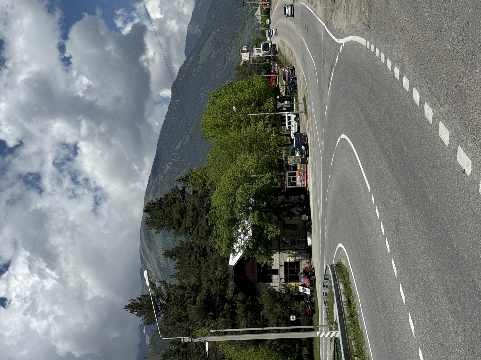 The image depicts a curved asphalt road with white dashed and solid lane markings, situated in the city of Teberda, Russia. The road curves to the right, leading towards a mountainous backdrop. On the left side of the road, a two-story building constructed of light-colored bricks is visible, with a dark-colored roof. Adjacent to the building, several red ATVs are parked. A streetlamp stands to the left of the building, and a traffic sign displaying "40" is mounted on its pole. Lush green trees surround the building and extend along the roadside. In the background, tall mountains are partially obscured by a cloudy sky. Several vehicles, including cars and a truck, are present on the road and its shoulder, suggesting ongoing activity. The scene appears to be a commercial or service area, possibly a rental or tour outpost, given the presence of ATVs and the building's layout. The environment is characterized by a mix of natural mountainous scenery and urban infrastructure.