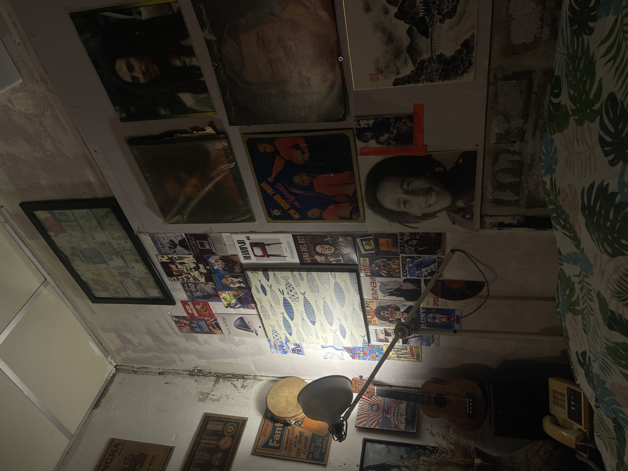 The image depicts a cluttered bedroom corner, likely in Chalong, Thailand. The walls are adorned with numerous posters and record covers, primarily featuring musicians like Bob Marley, along with other music memorabilia and artwork. A ukulele rests against a speaker, and a vintage-style telephone is visible on a nightstand. A floor lamp with an adjustable arm is positioned over the bed, which is covered with a tropical-patterned duvet. The overall impression is of a lived-in space with a strong emphasis on music and personal expression.
