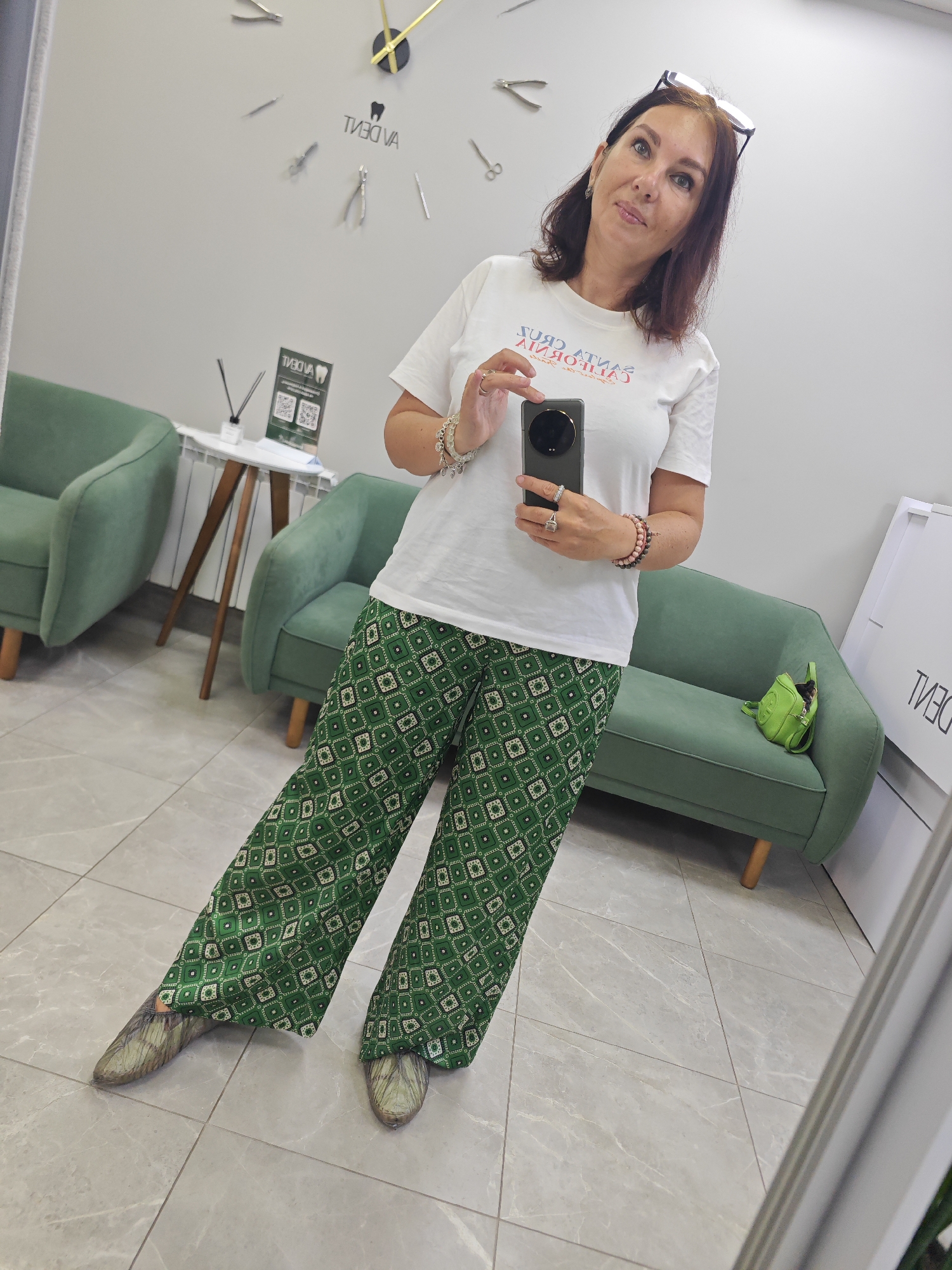 In the city of Lobnya, Russia, a woman stands in a bright room with pale gray walls and light gray tiled flooring. She is taking a mirror selfie, holding a dark-colored smartphone in her right hand. She is wearing a white t-shirt with text in red and blue, and green patterned wide-leg pants. Her shoes are a dull metallic green. The woman is wearing several bracelets on her left wrist and rings on her fingers. Her hair is dark brown and shoulder-length, and she is wearing glasses perched on top of her head.
Behind her, two mint green upholstered armchairs are visible. To the left of the chairs, a small white table with wooden legs holds a white vase with what appears to be a single dried reed. A small green bag is on the seat of a mint green sofa to her right. A large clock with golden hands and silver hour markers is mounted on the wall behind her, displaying the time as approximately 1:50. The clock face has the brand name "VADENT" in reverse lettering. A poster with the same brand name is also on the wall.