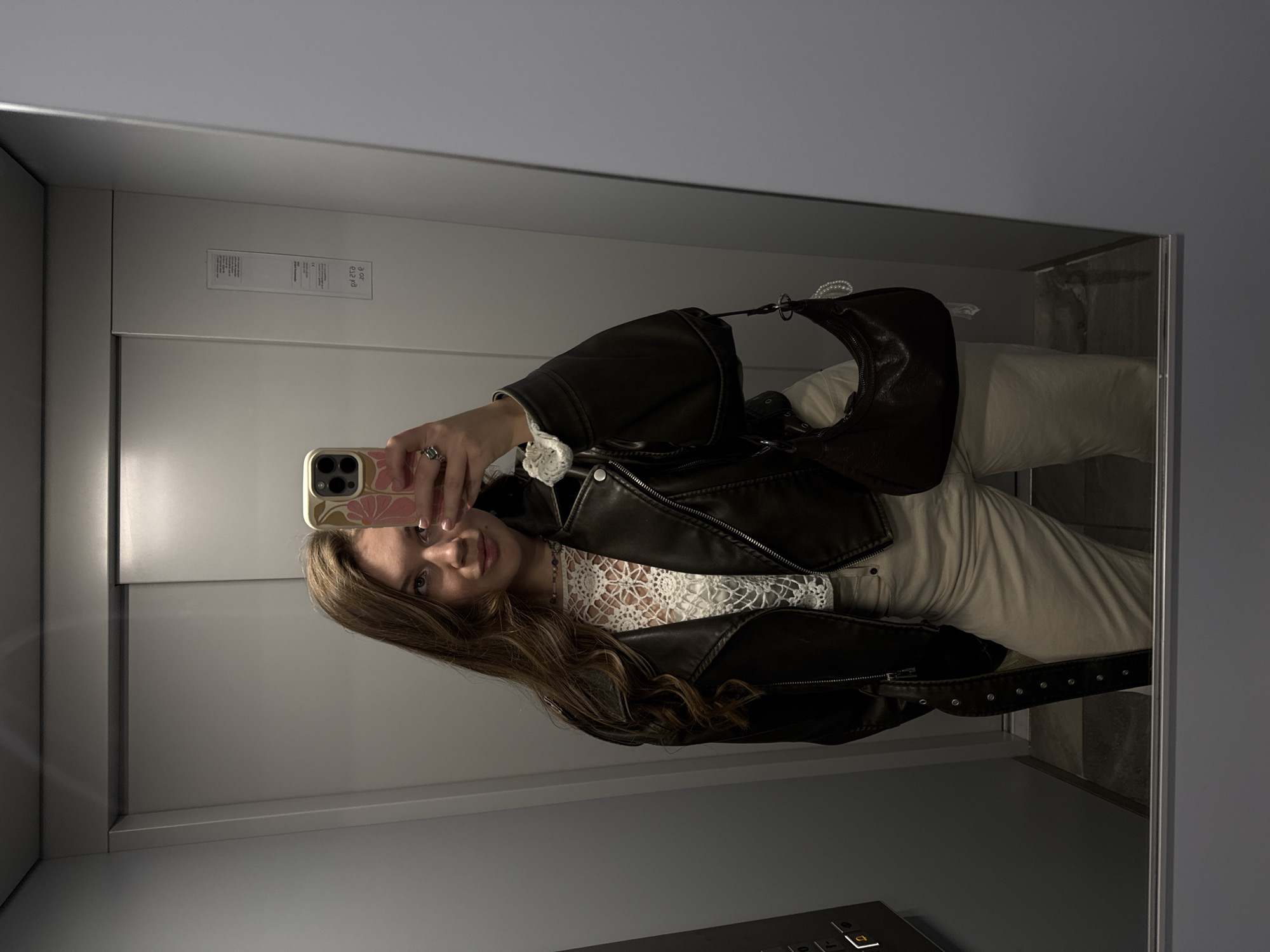 A young woman is taking a mirror selfie in an elevator. She is wearing a black leather jacket over a white lace top, beige pants, and carrying a brown purse. She is holding a smartphone with a floral case. The elevator has grey walls, with a panel of buttons visible in the lower right corner and labels/instructions on the wall behind the woman. There is a dirty reflection of foliage and/or stone on the right side of the mirror, suggesting the elevator doors face a natural landscape. The location details of Vozdovac Urban Municipality, Serbia are not confirmed in this image.