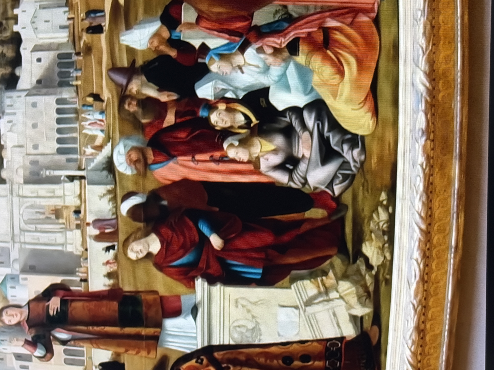 This is a detail from a painting depicting a group of people in what appears to be an outdoor setting with classical architecture in the background. In the foreground, several figures are gathered. A tall figure on the left stands draped in a dark brown and red robe over a white garment, with their left arm raised and hand pointing upwards. To their right, a woman in a red robe with blue undergarments is kneeling, looking upwards. Beside her, a group of individuals in various colored robes and headwear are seated or kneeling. One woman in a gray garment is sitting with her legs crossed, looking to the left. Another woman in a light blue garment with a blue head covering is looking upwards. To the right of these seated figures, a person in a bright orange and yellow robe is kneeling. In the middle ground, more figures are standing, some in discussion, and others observing. The background features large, pale buildings with arched windows and columns, suggesting a city or complex. The ground is depicted as a sandy or earthy color. The scene appears to be populated by individuals in historical or religious attire.