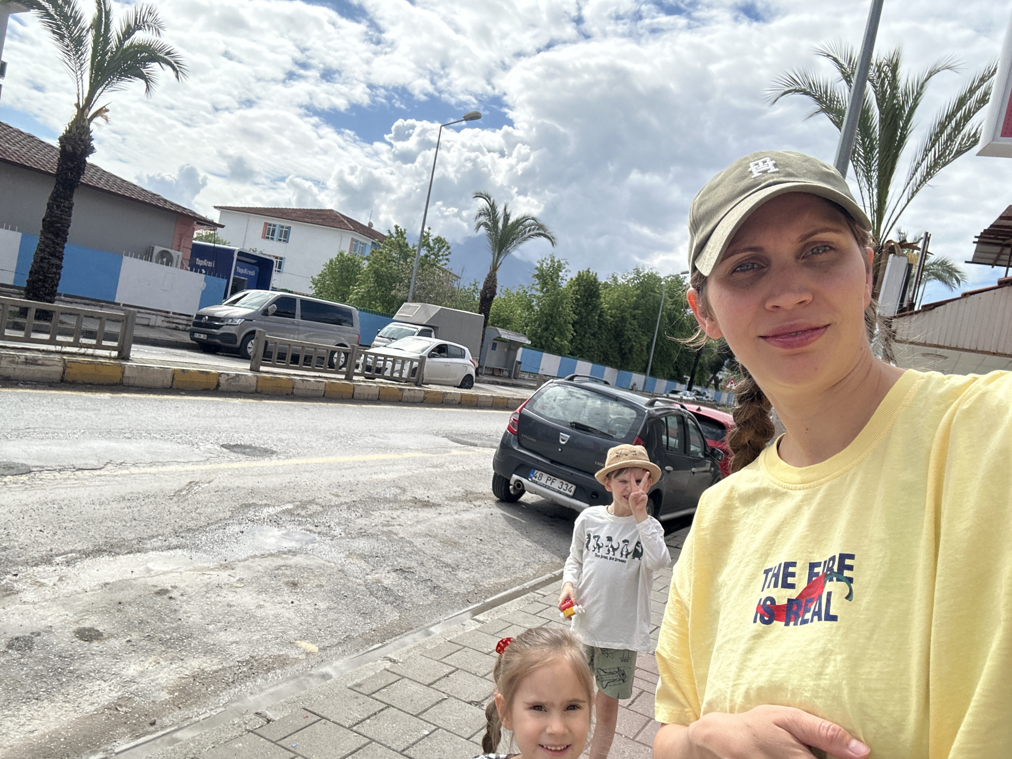 A selfie shows a woman with a child, likely in Fethiye, Turkey, given the Turkish license plate (48 PF 334) on a gray Dacia Sandero. The woman wears a yellow shirt with the phrase "The Fire is Real" and a baseball cap. In the background, there are parked cars including a Volkswagen minivan and a white hatchback, and buildings, with some palm trees. A child wearing a sunhat stands partially behind her, flashing a peace sign, and a younger child is partially in view towards the bottom left corner. The setting appears to be an urban street on a cloudy day.