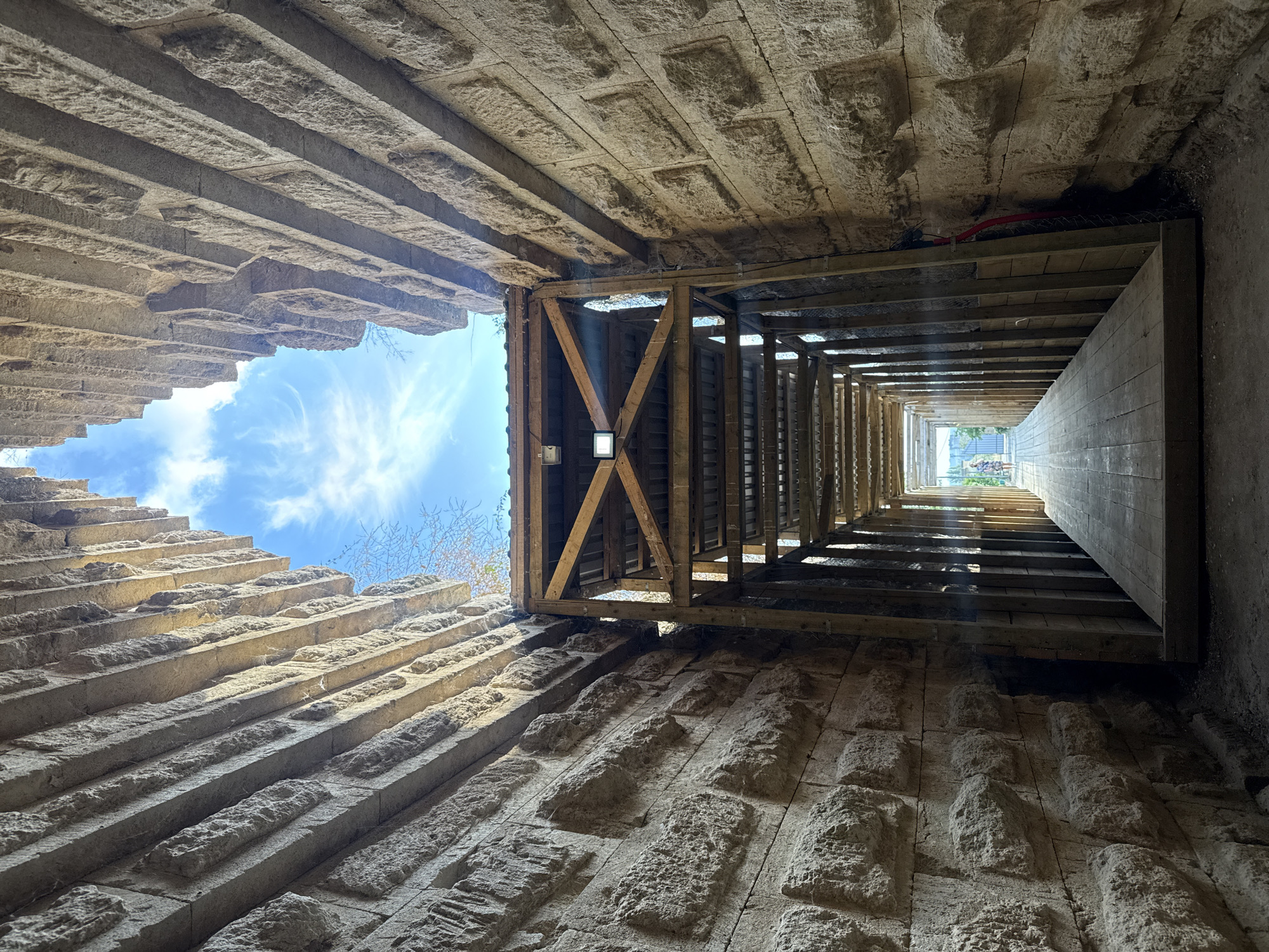 This is a ground-level, eye-level, vertical shot of an archway leading to an open sky, located in Kerch, Ukraine. The archway is constructed from large, rough-hewn sandstone blocks. The pathway within the archway is covered by a wooden structure, which appears to be under construction or renovation. The wooden structure has a sloped roof made of corrugated metal, supported by wooden beams. A spotlight is attached to the wooden framework. The pathway itself is made of wooden planks. In the distance, at the end of the pathway, a woman in a blue dress and a man in a purple shirt are visible, approaching the exit of the archway. The sky is blue with scattered white clouds.