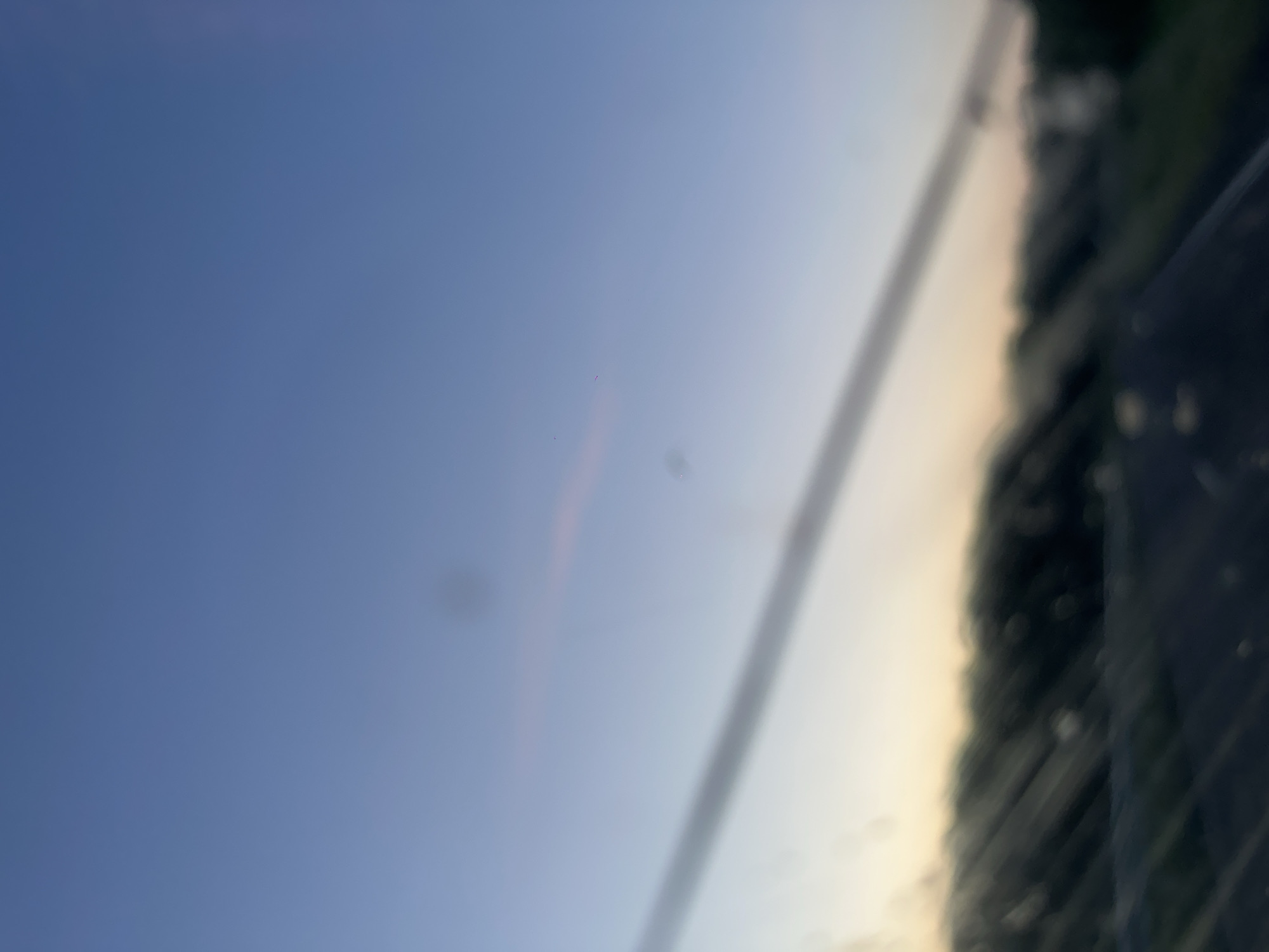 The image shows a blurred view of a landscape at dusk, possibly taken from a vehicle. The sky is a gradient of blue and pale yellow, with a faint pinkish hue near the horizon. A dark, diagonal line, possibly a power line or a reflection on a window, stretches across the upper portion of the image. Below this, a blurry line of trees or foliage is visible, with a dark road or ground in the foreground. The overall impression is one of a tranquil, transitional moment at the end of the day. The location in Krapivinskiy, Russia, is not visually discernible from the image itself; this information is external to the visual content.