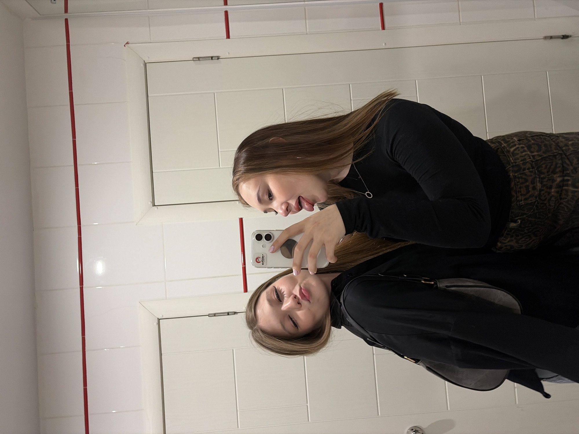 The image shows a young woman taking a selfie in a public restroom. The restroom features white tiled walls with red vertical lines along the corners. The woman is dressed in a black long-sleeved shirt and leopard print pants. She is holding a white iPhone, with the screen facing toward the mirror. She is posing in front of the mirror, and her reflection is visible. Based on the information available, the location is Chaikovsky, Russia.