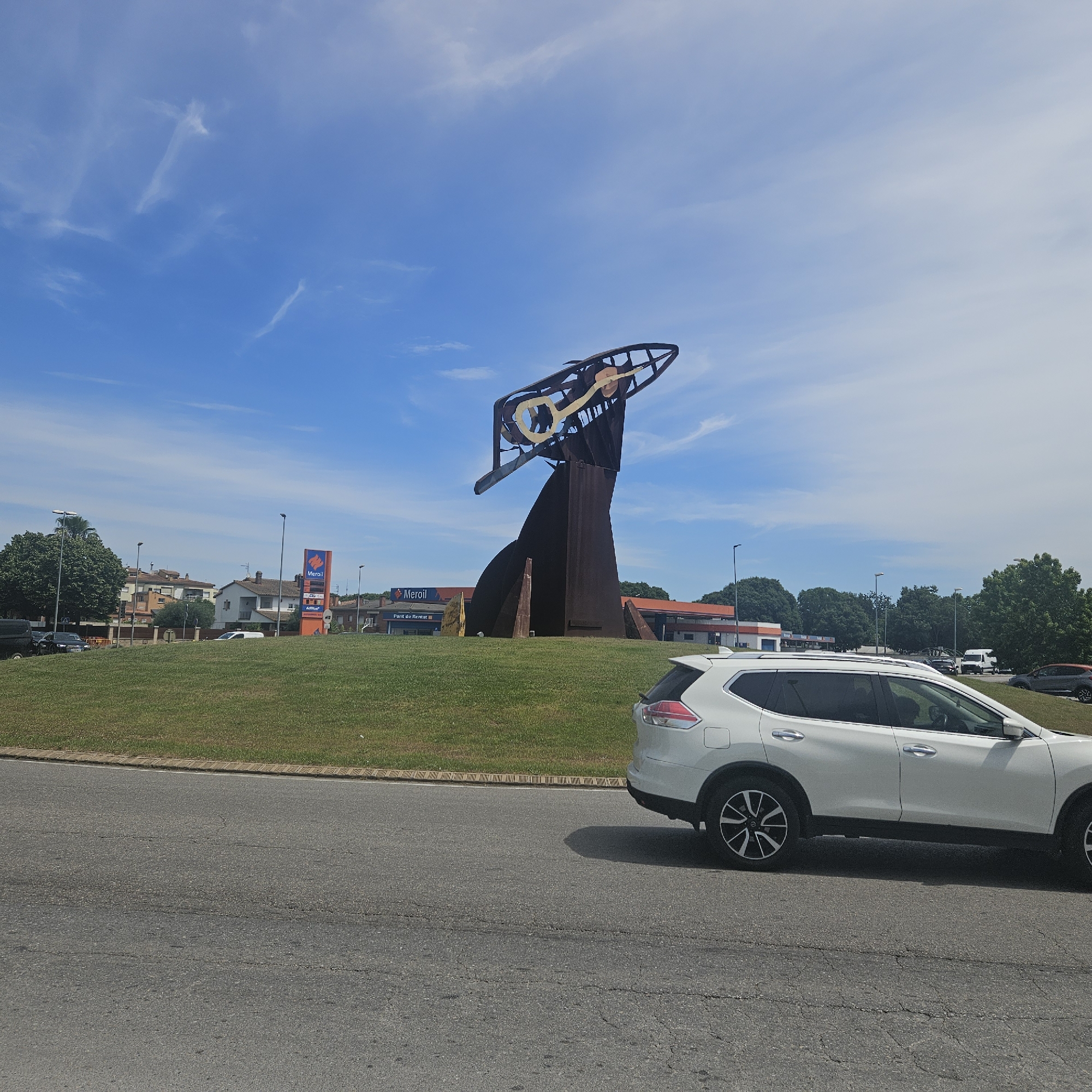 This daylight image depicts a roundabout on Ronda de Sant Antoni / Carretera de Girona in Salt, Girona, Spain, under a clear blue sky with wispy white clouds. The foreground features dark gray asphalt road exhibiting minor cracks, adjacent to a textured brown curb and a well-maintained green grassy median.
At the center of the grassy median stands a large, abstract sculpture, known as "El Llaurador" (The Harvester). It is constructed from dark, weathered metal, likely steel, and features an upward-curving, open framework. A prominent stylized golden-yellow key or wrench-like shape is integrated into the upper portion of the sculpture.
Behind the sculpture, a Meroil gas station is visible, identified by a tall, orange sign displaying "Meroil" and "Pont de Rentat" in white text. Further back, two-to-three-story residential buildings with light-colored facades and terracotta roofs are partially obscured by trees. Multiple tall, dark streetlights are scattered across the scene.
A white Nissan Rogue SUV is prominently positioned on the asphalt in the lower right, facing left. Other vehicles, including a dark SUV and a white van, are visible in the mid-ground. Lush green foliage from various trees, including some broadleaf species and a distant palm, frames the periphery of the urban landscape. Bright sunlight casts distinct shadows, particularly from the white SUV onto the road.