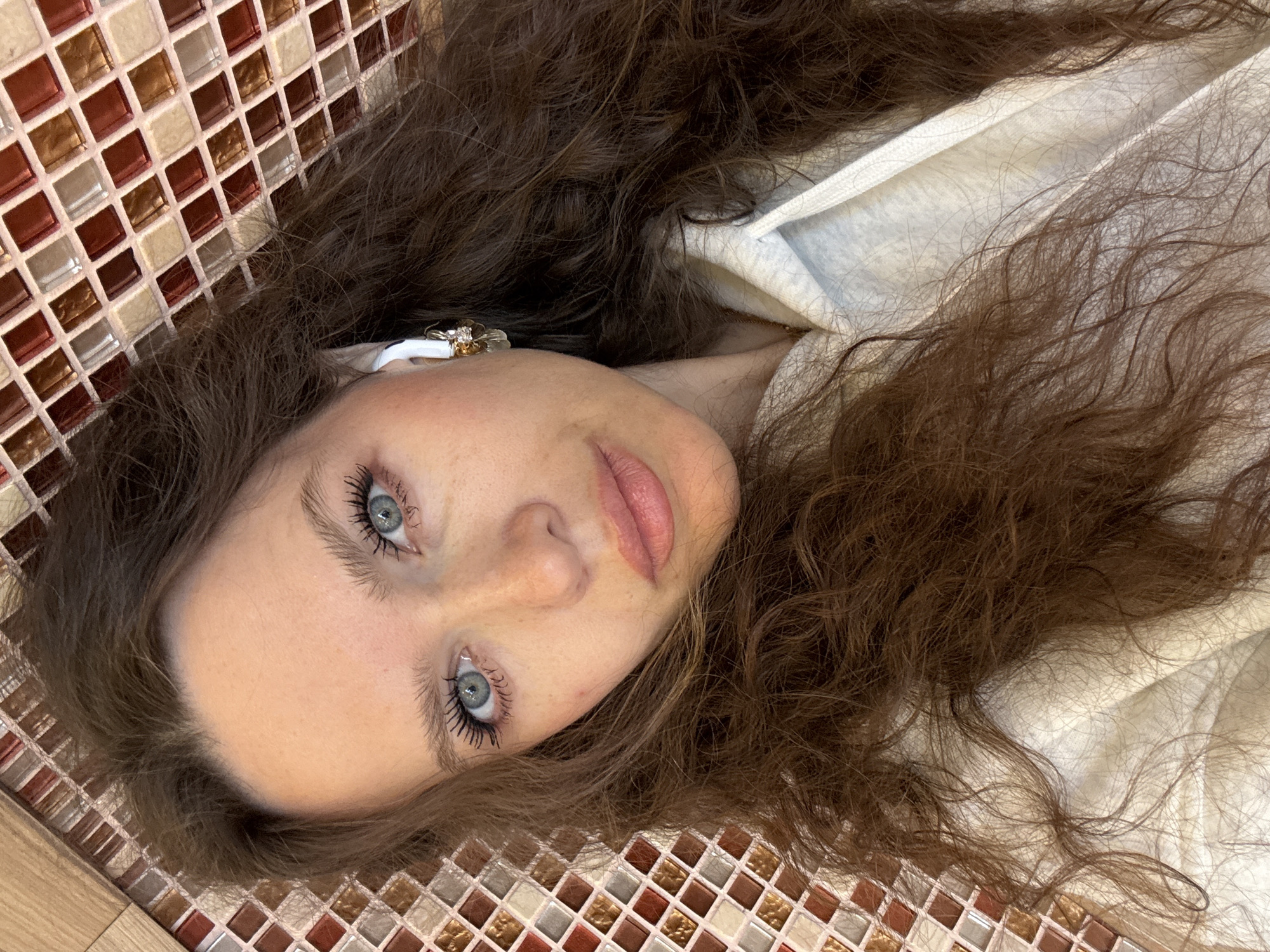 A fair-skinned woman with long, wavy brunette hair and light blue eyes lies on her back, presumably indoors. She is wearing a white hooded garment. Her head rests on a surface composed of small, mosaic tiles of varying shades of brown, beige, and white, and a wooden surface is visible in the lower left corner. The environment is nondescript. There is no indication that the location is Moscow, Russia.