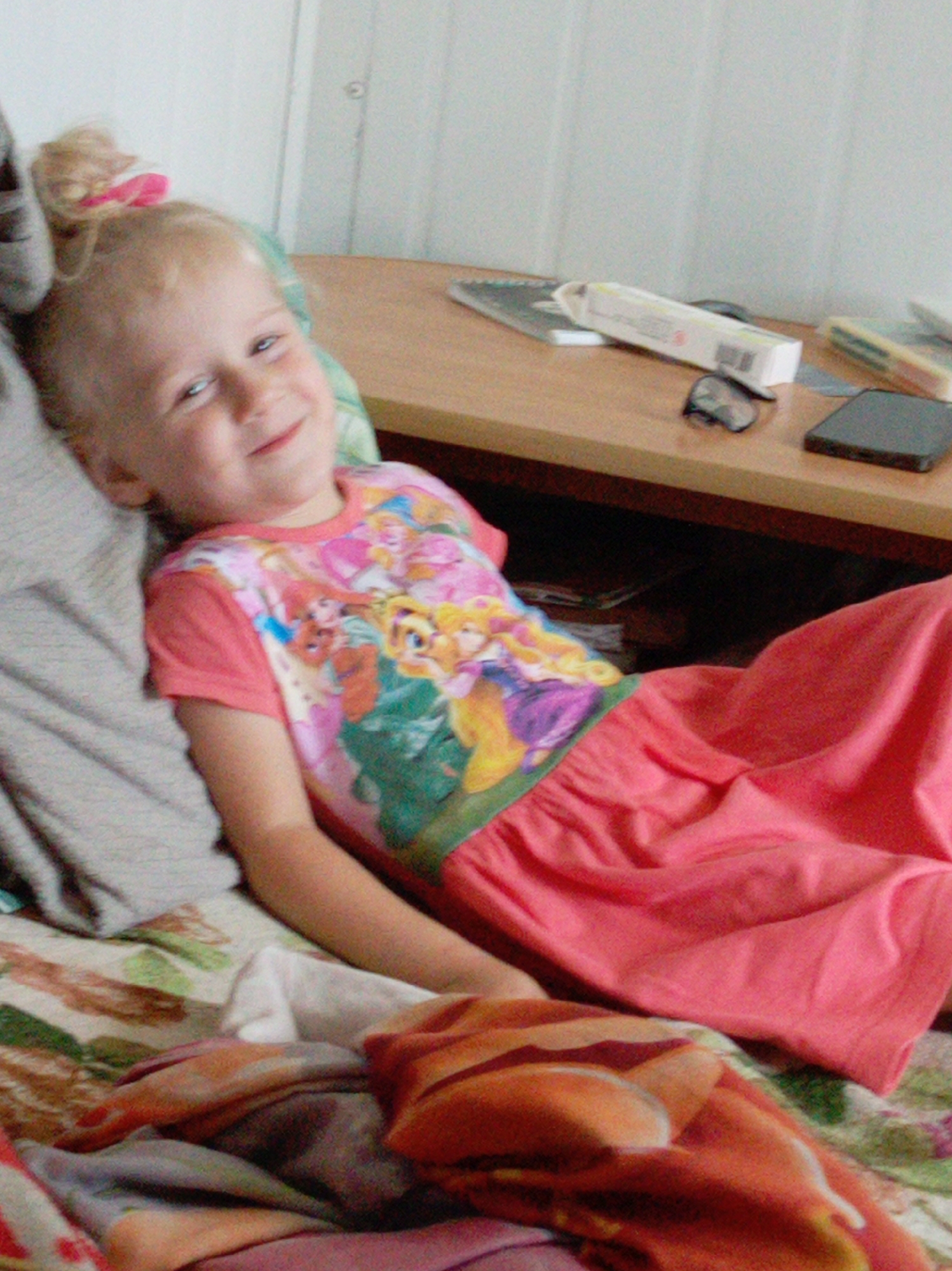 A young child, appearing to be a girl, is lying on a bed. She has blonde hair styled in a ponytail with a pink accessory. She is wearing a pink t-shirt with a print of cartoon princesses. The child is looking towards the viewer with a slight smile. She is lying on a bed with a patterned duvet. In the background, a wooden table is visible with several objects on it, including a white rectangular box, a pair of glasses, and a dark rectangular object, possibly a phone. The room appears to be indoors, with a white wall in the background. The lighting suggests it is daytime.