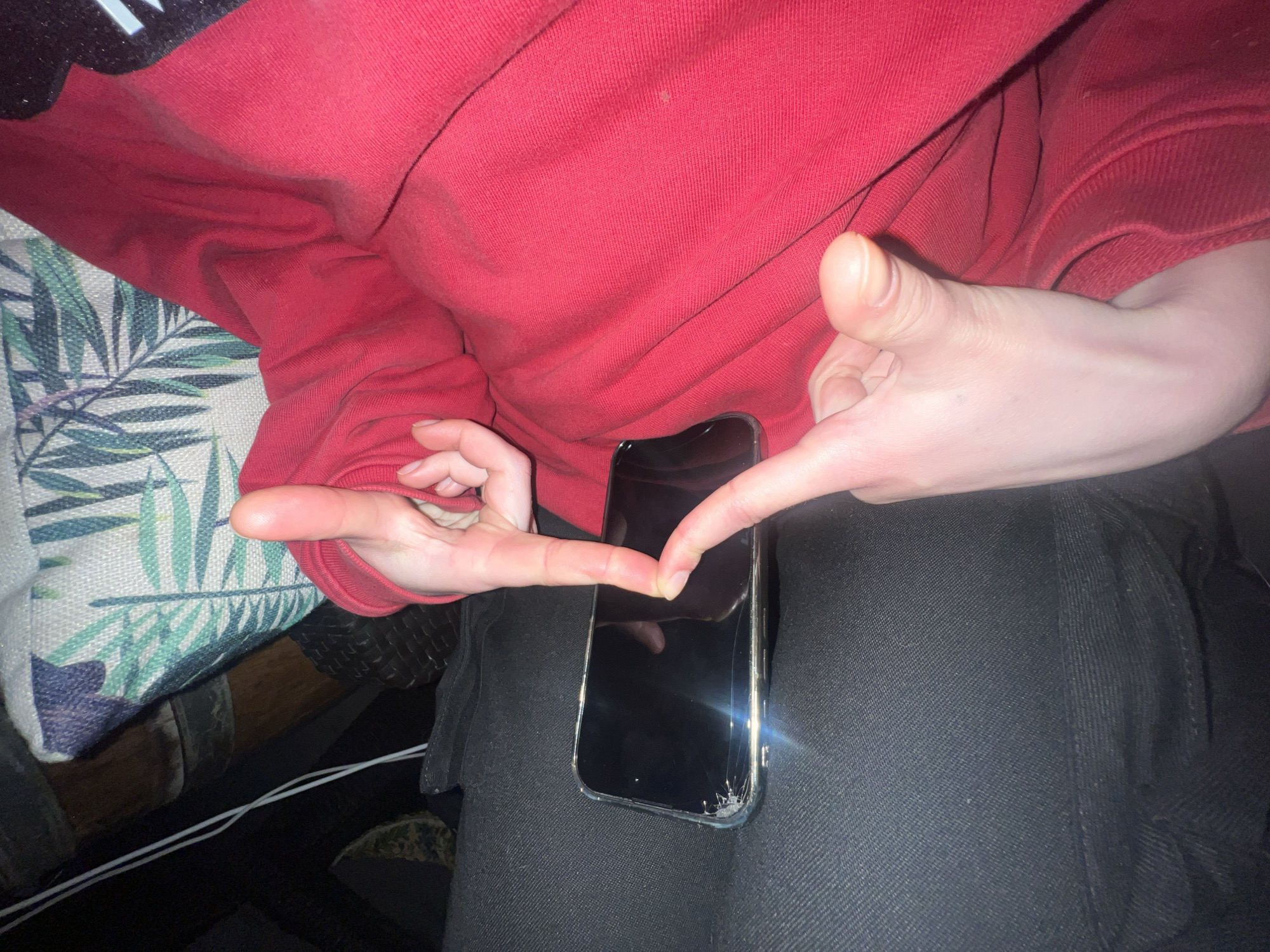 A person wearing a red sweatshirt and black pants is sitting on a sofa. Their hands are positioned around a black smartphone with a cracked screen. The person is making a "V" sign with their fingers around the phone. The sofa has a pillow with a green leaf pattern. There is no indication of the location being Sochi, Russia.
