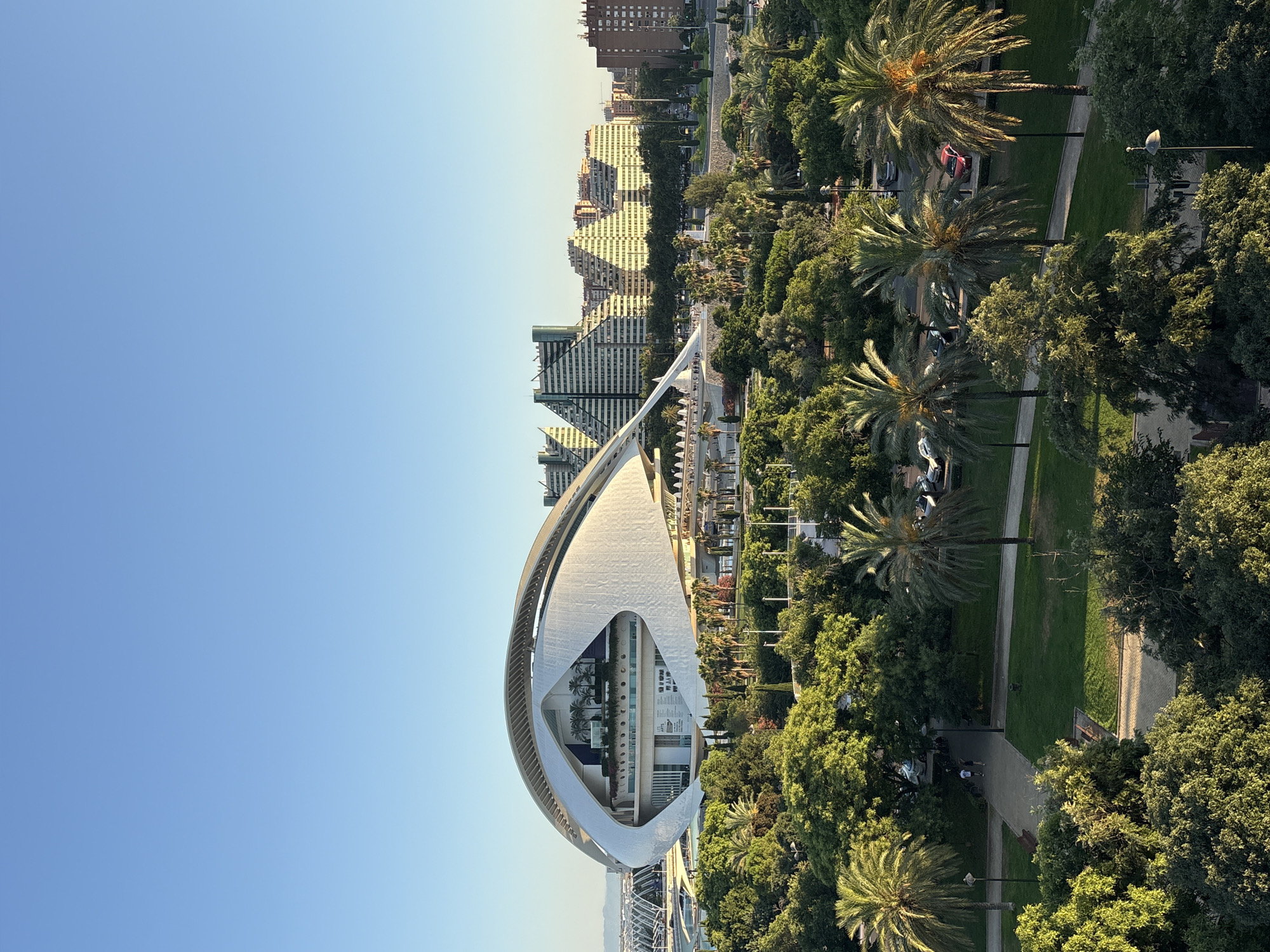 This is a high-angle, full-shot photograph of the City of Arts and Sciences in Valencia, Spain, during daylight. The sky is clear and blue. The foreground consists of lush green trees and palm trees, with a pathway and grass visible in a park area. In the midground, the distinct architectural structure of the L'Hemisfèric, a planetarium and IMAX cinema, dominates the scene. It is a modern, white building with an eye-shaped, parabolic roof that reflects in a nearby pool. Behind L'Hemisfèric, several other modern buildings with geometric designs and pyramidal shapes can be seen, characteristic of the Santiago Calatrava-designed complex. Further in the background, residential buildings and a hint of the distant landscape are visible under the bright sky. The overall impression is of a vibrant, modern urban landscape integrated with green spaces.