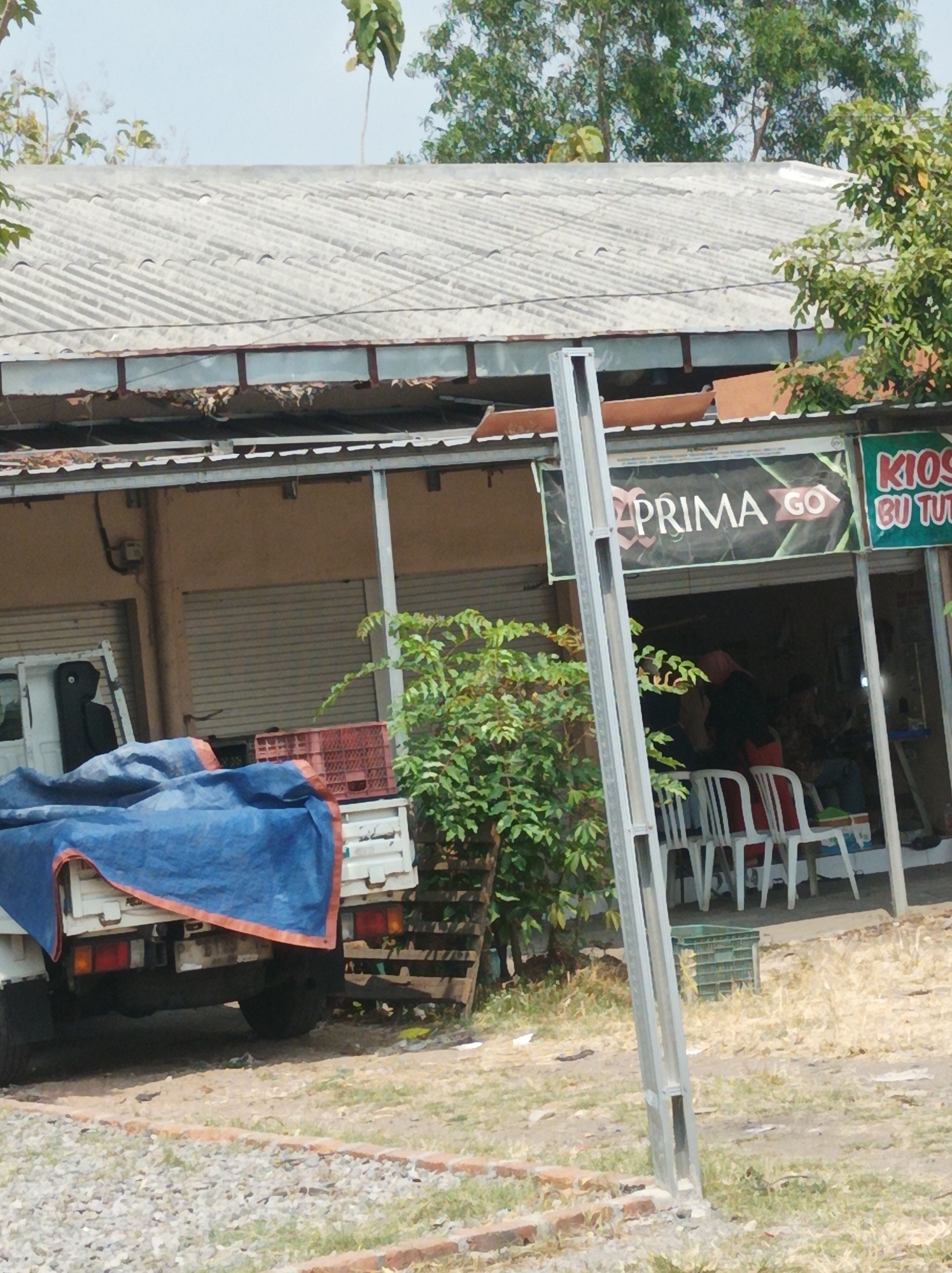 The image depicts an outdoor scene in Nganjuk, Indonesia, featuring a building with a corrugated metal roof and an open-fronted section that functions as a kiosk or shop. A white truck, partially covered by a blue tarp with orange trim, is parked in front of the building. Red plastic crates are visible on the truck bed. Inside the open area of the building, several chairs are arranged, and people are seated. A banner with the text "PRIMA GO" is prominently displayed above the entrance, and another sign with the text "KIOS BU TUTI" is visible to the right. The ground is covered with dry grass and gravel. A metal pole stands in the foreground, and trees and vegetation surround the scene.
