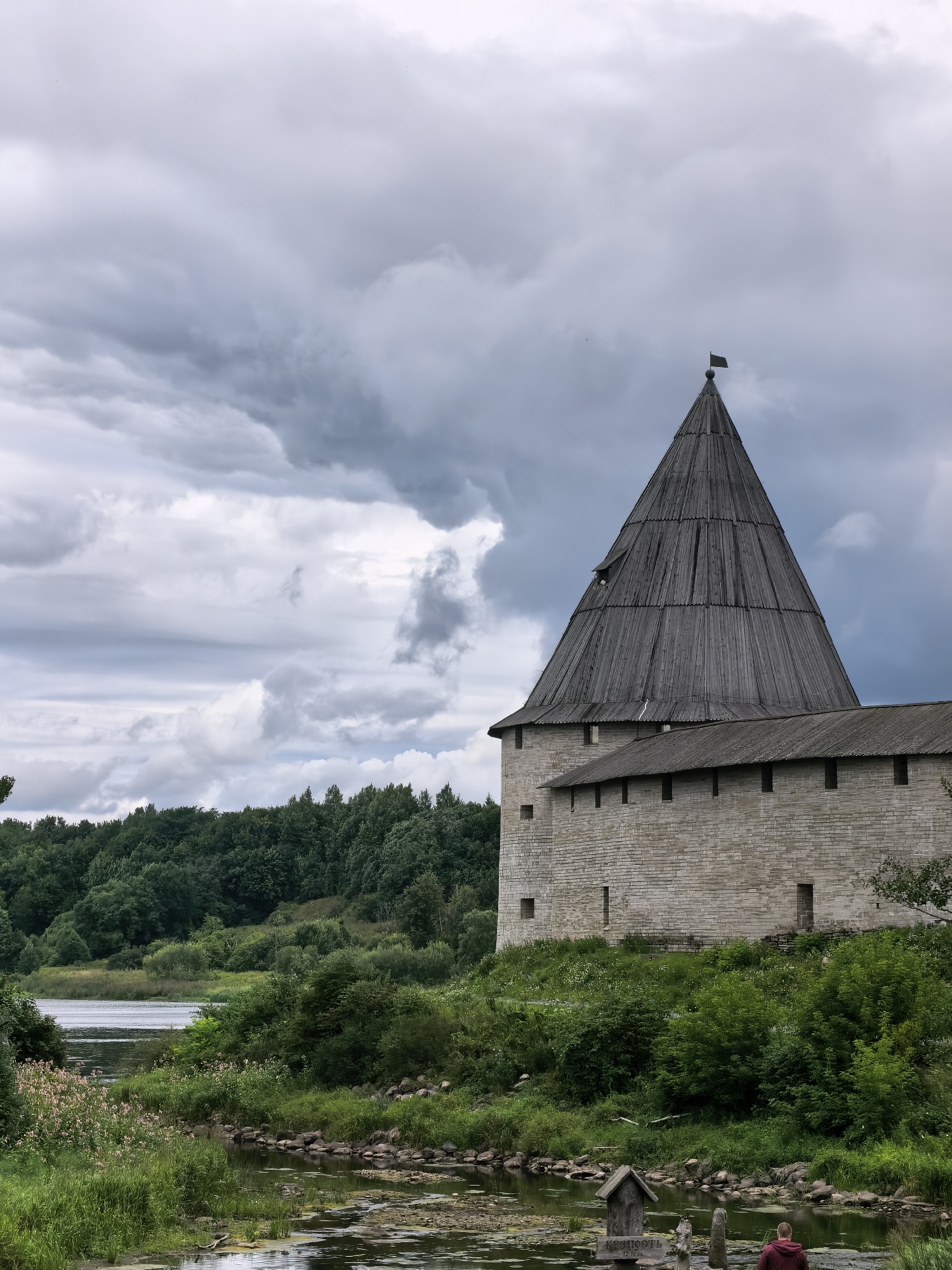This image depicts the Staraya Ladoga Fortress in Staraya Ladoga, Russia, a historical landmark on the Volkhov River. The fortress features a prominent stone tower with a conical wooden roof, topped by a dark flag. The stone walls of the fortress are constructed from large, roughly hewn blocks. Surrounding the fortress is lush green vegetation, including dense trees on a distant hill and grass and shrubs closer to the foreground. A body of water, likely the Volkhov River or a connected channel, is visible in the lower left portion of the frame, with reeds and scattered rocks along its edge. In the immediate foreground, a small wooden structure is visible near the water's edge, with two stone posts beside it. A person wearing a maroon hooded sweatshirt stands with their back to the camera, facing the water near the wooden structure. The sky above is overcast with dramatic, dark grey clouds.