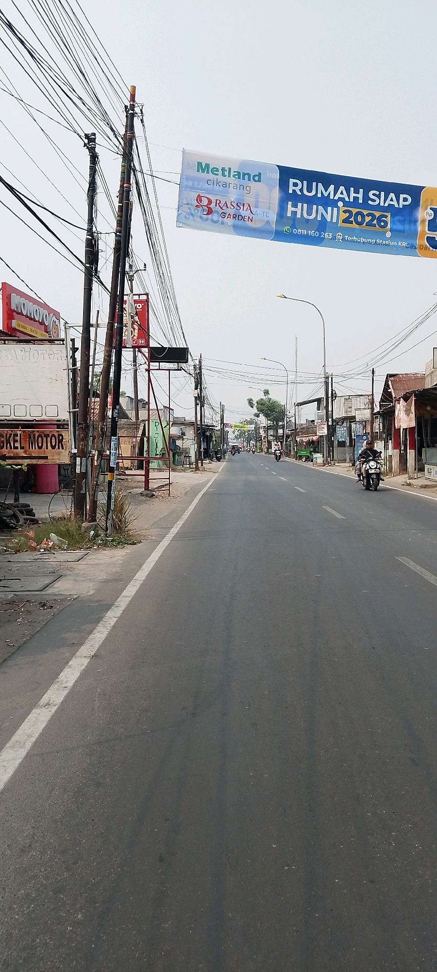 A street in Satriamekar, Indonesia, is depicted under a cloudy sky. The road is paved and has a white lane marker on the left side. Several utility poles with numerous wires are visible along the street. A banner is stretched across two poles, displaying text that includes "Metland Cikarang" and "RUMAH SIAP HUNI 2026." On the left side of the road, there are businesses with signs, including one reading "MOMOYO." Further down the street, several people are riding motorcycles. The overall environment appears to be a commercial or mixed-use area with some signs of development.