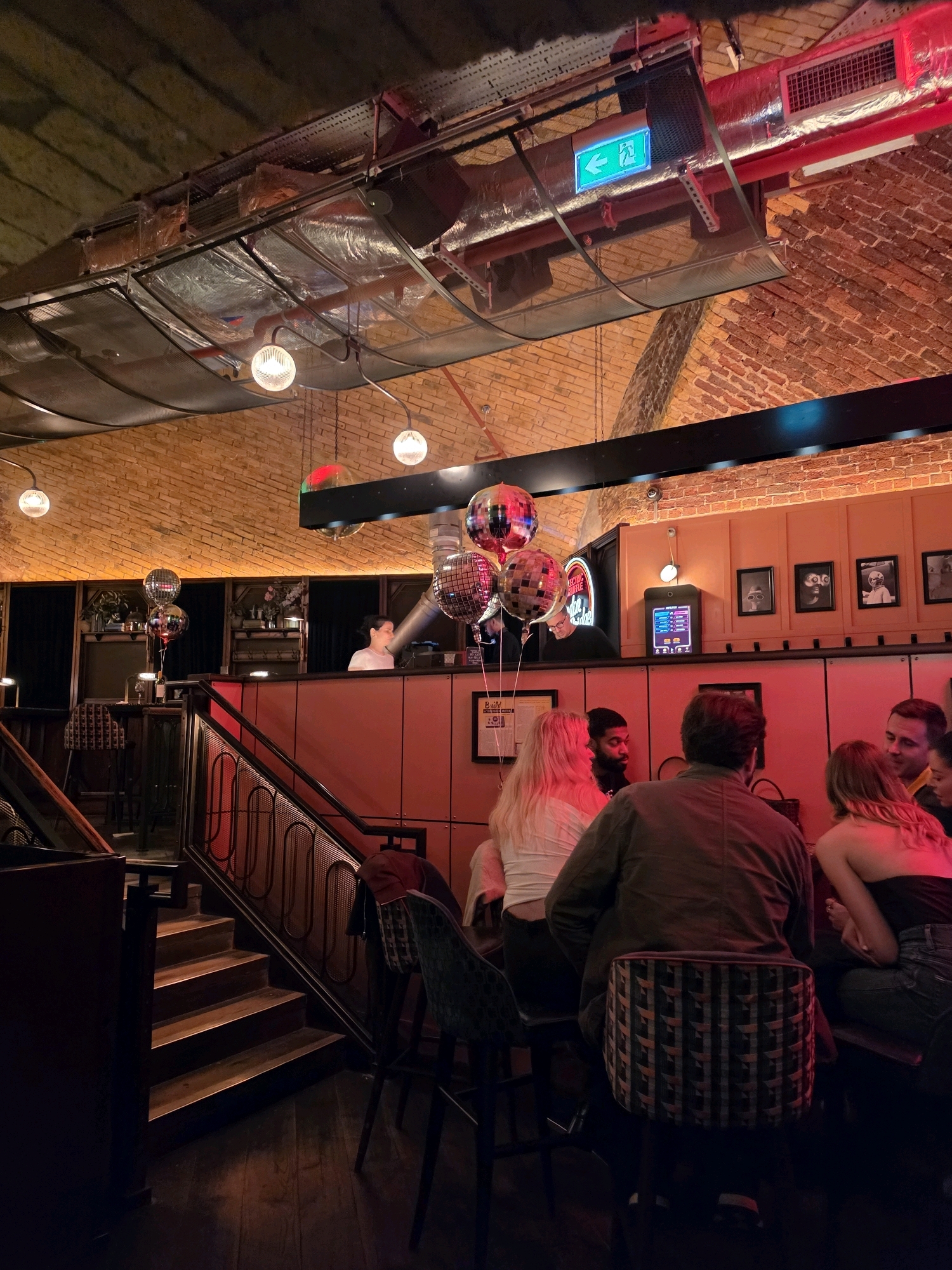 The image depicts the interior of a dimly lit bar or club in London, United Kingdom. The scene is characterized by warm, reddish lighting, exposed brick walls, and industrial-style ductwork visible on the ceiling. Several groups of people are seated at tables and a bar area, some engaged in conversation. The ambiance suggests a social gathering or entertainment venue.
In the foreground, a section of wooden stairs leads up to a raised bar area. Behind the bar, a woman is visible working. Several disco balls hang from the ceiling, adding a festive touch to the environment. The overall impression is of a modern, stylish establishment with a relaxed atmosphere.