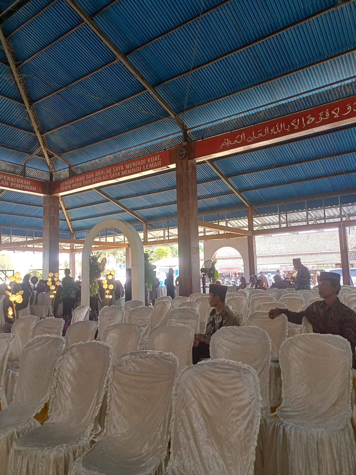 The image shows a wide-angle view of an outdoor event space, likely a community hall or pavilion, in Wanadadi, Indonesia. The structure has a corrugated blue roof supported by metal beams and wooden pillars. Red banners with Arabic calligraphy and Indonesian text are visible, one reading "PERATURAN DAN PIMPINAN" and another stating "DENGAN IMAN DAN AKHLAQ SAYA MENJADI KUAT, TANPA IMAN DAN AKHLAQ SAYA MENJADI LEMAH."
The foreground is filled with rows of white-covered chairs, many of them empty. Several people are scattered throughout the seating area and in the background. In the middle ground, an arched doorway leads to another area where more people are gathered, and decorative lamps with yellow lights are placed among the chairs. Two men in traditional Islamic attire, including peci hats, are seated in the foreground, looking towards the left and center of the frame. Another man in similar attire is visible further back, near the right edge of the frame, with his back to the camera. The overall scene suggests a gathering, possibly a religious or community event, under a covered outdoor structure.
