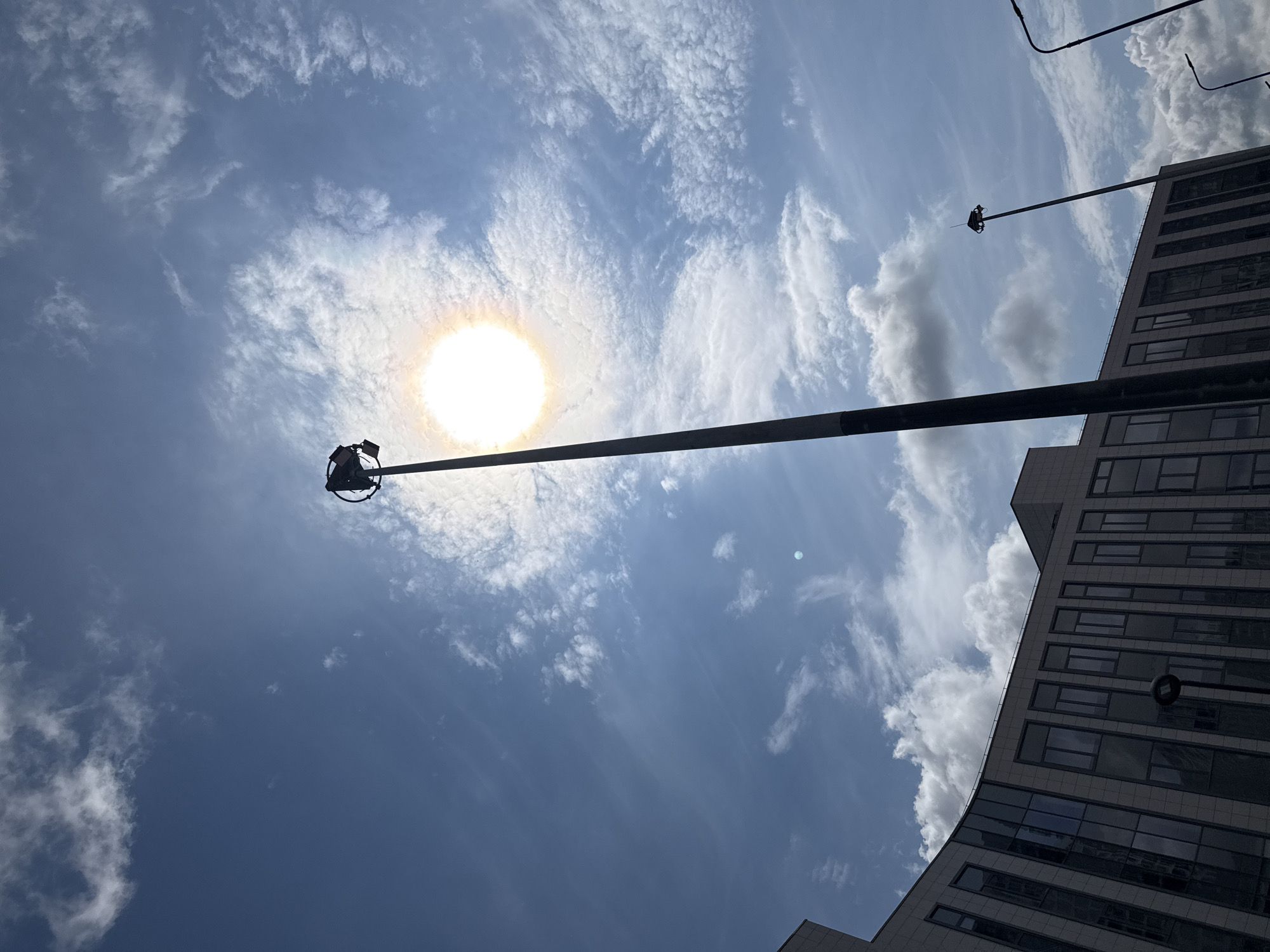 The image depicts a bright, sunny day with a clear blue sky dotted with wispy clouds. A tall, dark lamppost dominates the center of the frame, extending upwards towards the sun, which is intensely bright and partially obscured by clouds, creating a halo effect. Atop the lamppost, a circular fixture with several lights is visible. In the background, the facade of a modern building with many windows and a curved design is seen. To the right, another lamppost with a curved arm and a light fixture is partially visible. The scene appears to be an outdoor urban environment, possibly in the city of Lyubertsy, Russia, given the context. There are no people or animals visible in the image.