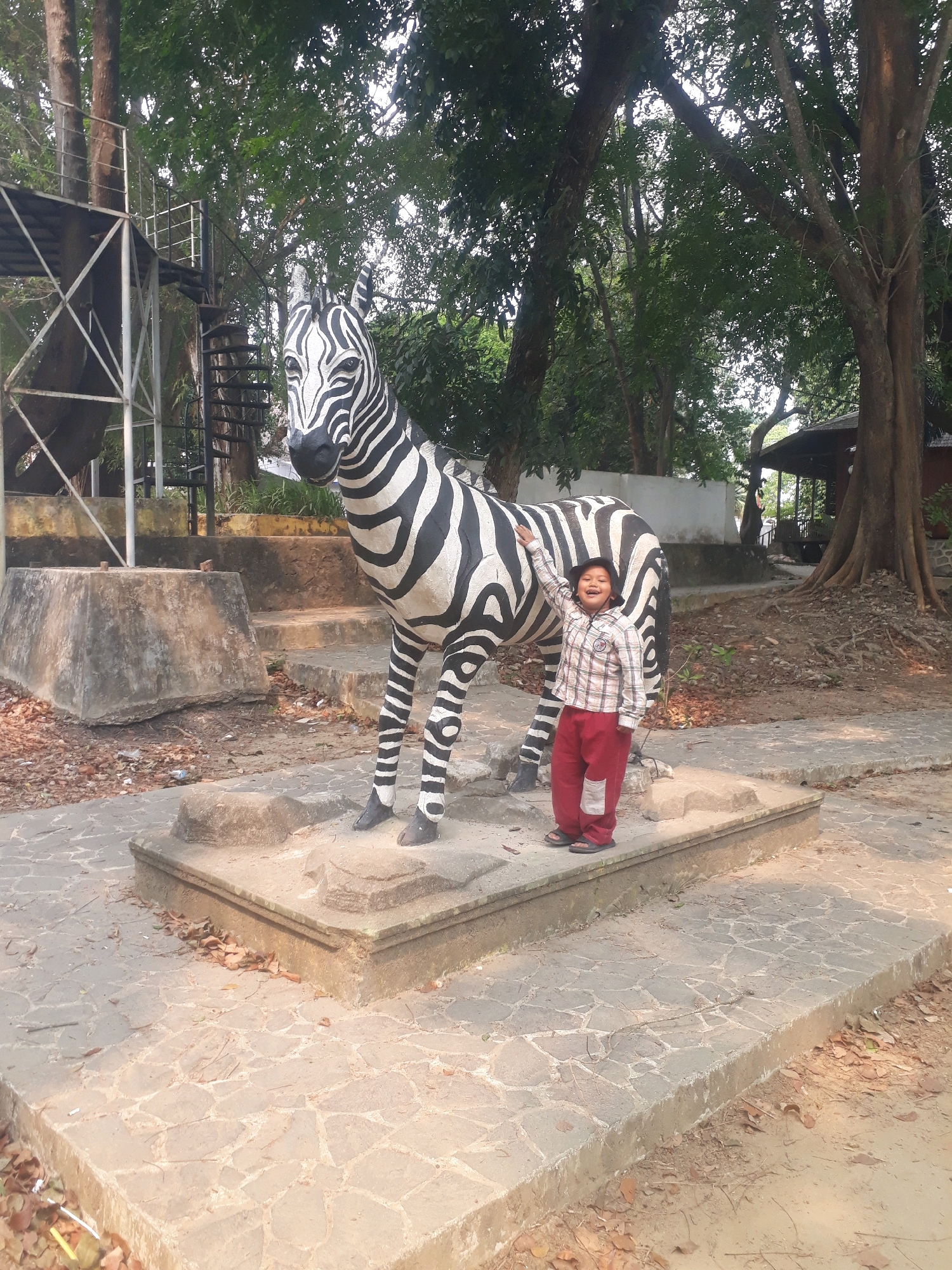 A young boy wearing a plaid shirt and red pants stands to the right of a large zebra statue in a park setting. The boy has his left arm raised, touching the zebra's side, and appears to be smiling. The zebra statue is black and white, with detailed stripes. It is positioned on a concrete platform with stone paving in front of it. The background features lush green trees, suggesting an outdoor, possibly tropical environment. To the left of the zebra, a metal structure with stairs is visible, possibly a viewing platform or part of an attraction. The ground is covered with fallen leaves and some debris. The scene is well-lit, indicating daytime. The location is the Pangkalpinang City Park, Bangka Belitung Islands Province, Indonesia.