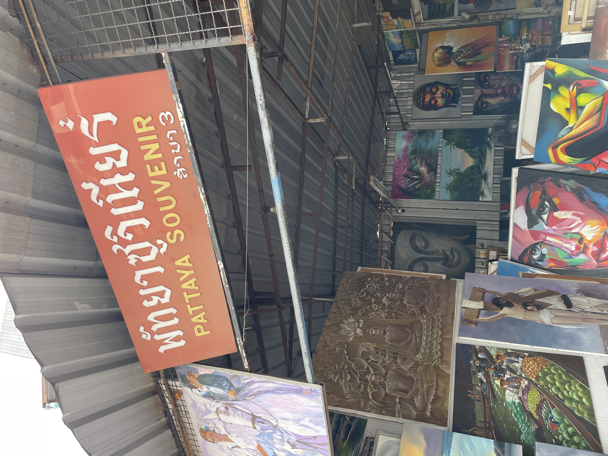 The image captures a street-side art stall in Pattaya City, Thailand, under a corrugated metal roof structure. A prominent orange sign with white and yellow lettering reads "พัทยา ซูวิเนียร์" and "PATTAYA SOUVENIR" with Thai characters below. The stall displays a variety of paintings and artwork.
In the foreground, several canvases are stacked, including a large painting of a woman's face with vibrant, abstract colors, and another featuring a figure on a cross with a supporting figure. To the left, a painting depicts people in boats filled with fruit in a canal.
Above, more artwork is visible, including a large bas-relief style carving of Buddha figures surrounded by lotus flowers, and paintings of Buddhist imagery, landscapes with palm trees, and a scene with pink blooming trees. A portrait of a Buddha face and a painting of a woman in a headscarf are also displayed.
The stall appears to be outdoors, with metal beams and wires supporting the roof and holding up the artwork. The overall impression is a bustling market stall selling souvenirs and art in a tropical setting.