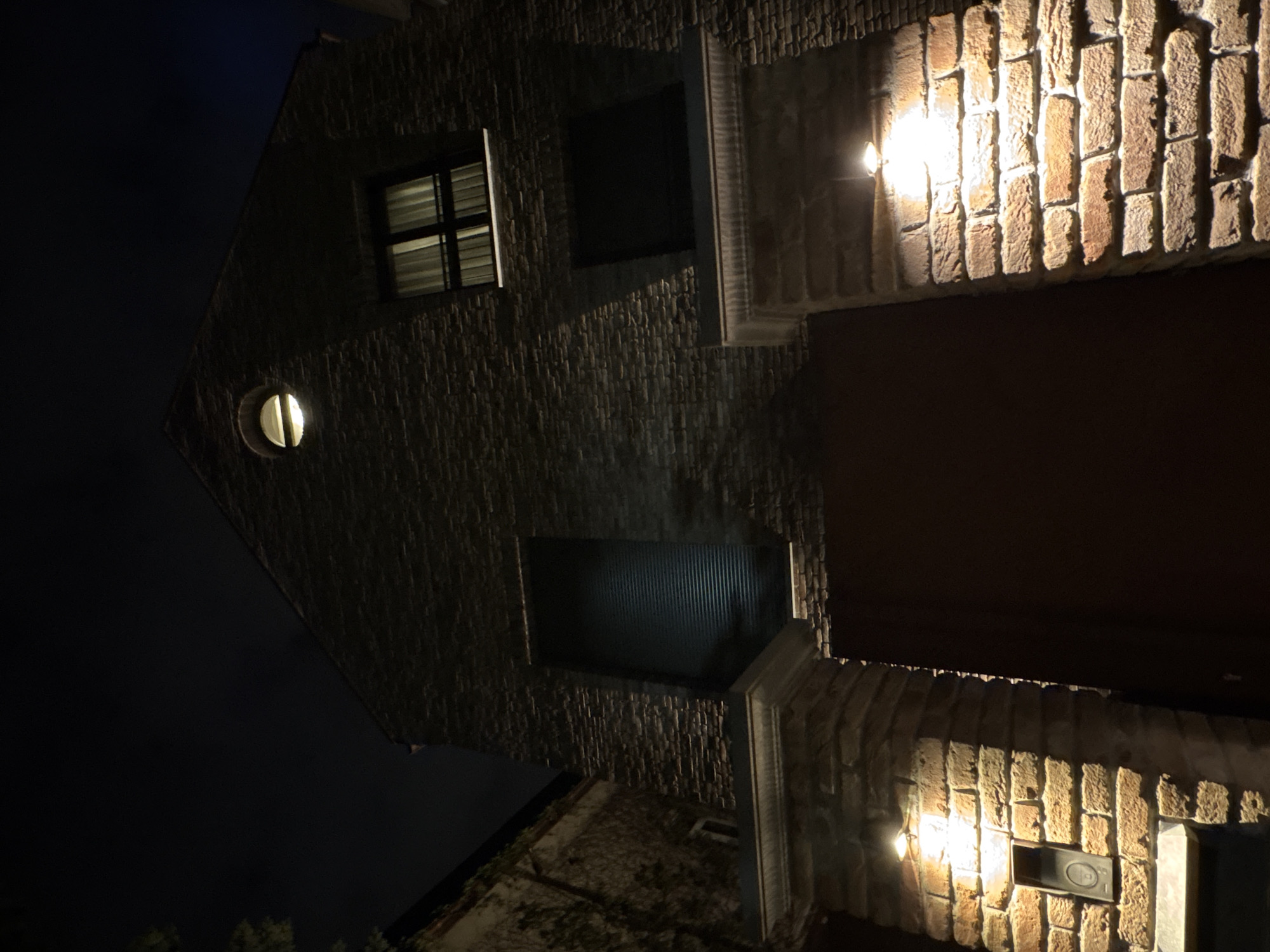 Exterior night shot of a building with brickwork. Features include two lit windows, a round lit window, and exterior light fixtures. The location is likely Sofia, Bulgaria.