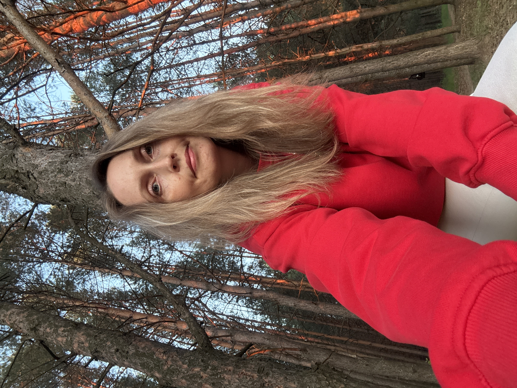 A woman with blonde hair wearing a red sweatshirt and white pants is taking a selfie in a forest. The trees are tall and thin, with a canopy of branches and needles. The sun is setting, casting a warm glow on the trees. The woman is smiling and looking at the camera. The location is identified as Chaikovsky, Russia.