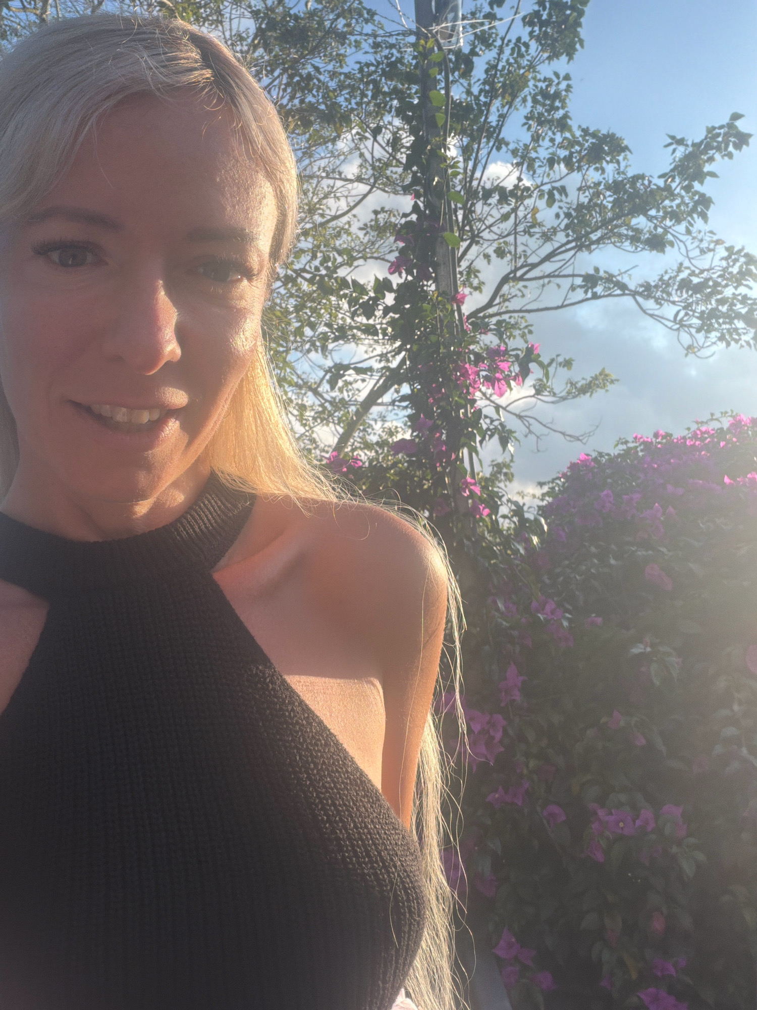 A woman with blonde hair and a black top is taking a selfie outdoors. She is smiling and looking at the camera. Behind her, there are trees, bushes with purple flowers, and a blue sky with clouds. The sun is shining brightly, casting a warm glow on her face and shoulders. The location appears to be in a tropical or subtropical climate, consistent with Xuân Thành, Vietnam.