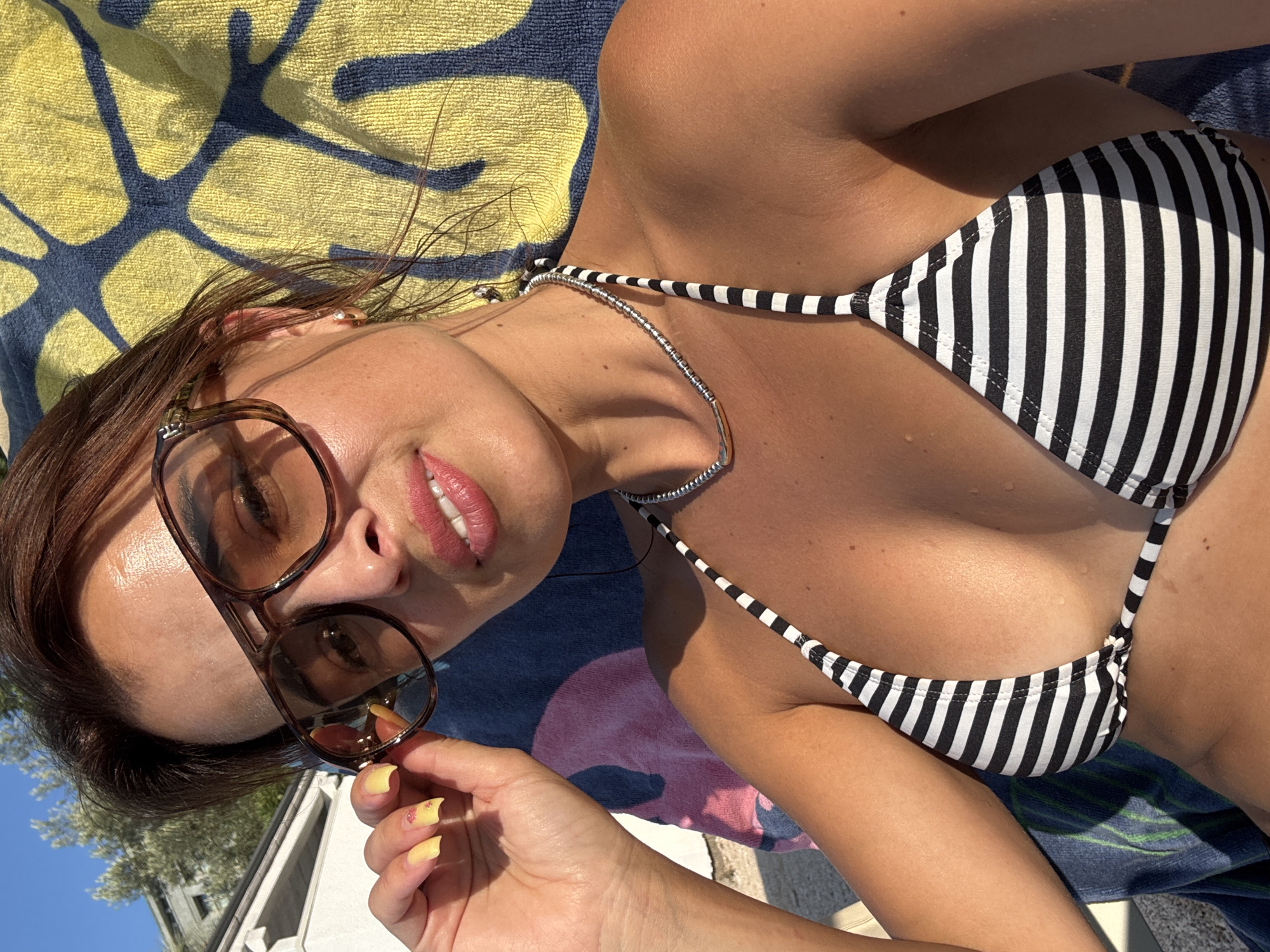 A woman with brown hair and brown eyes is captured in a close-up selfie, wearing large brown sunglasses and a black and white striped bikini top. Her left hand is raised, adjusting the sunglasses. She has on light pink lipstick and yellow nail polish. A silver necklace adorns her neck. The woman is sitting outdoors, leaning against a towel with a yellow and blue abstract pattern. The background suggests a sunny day with hints of a building and trees.