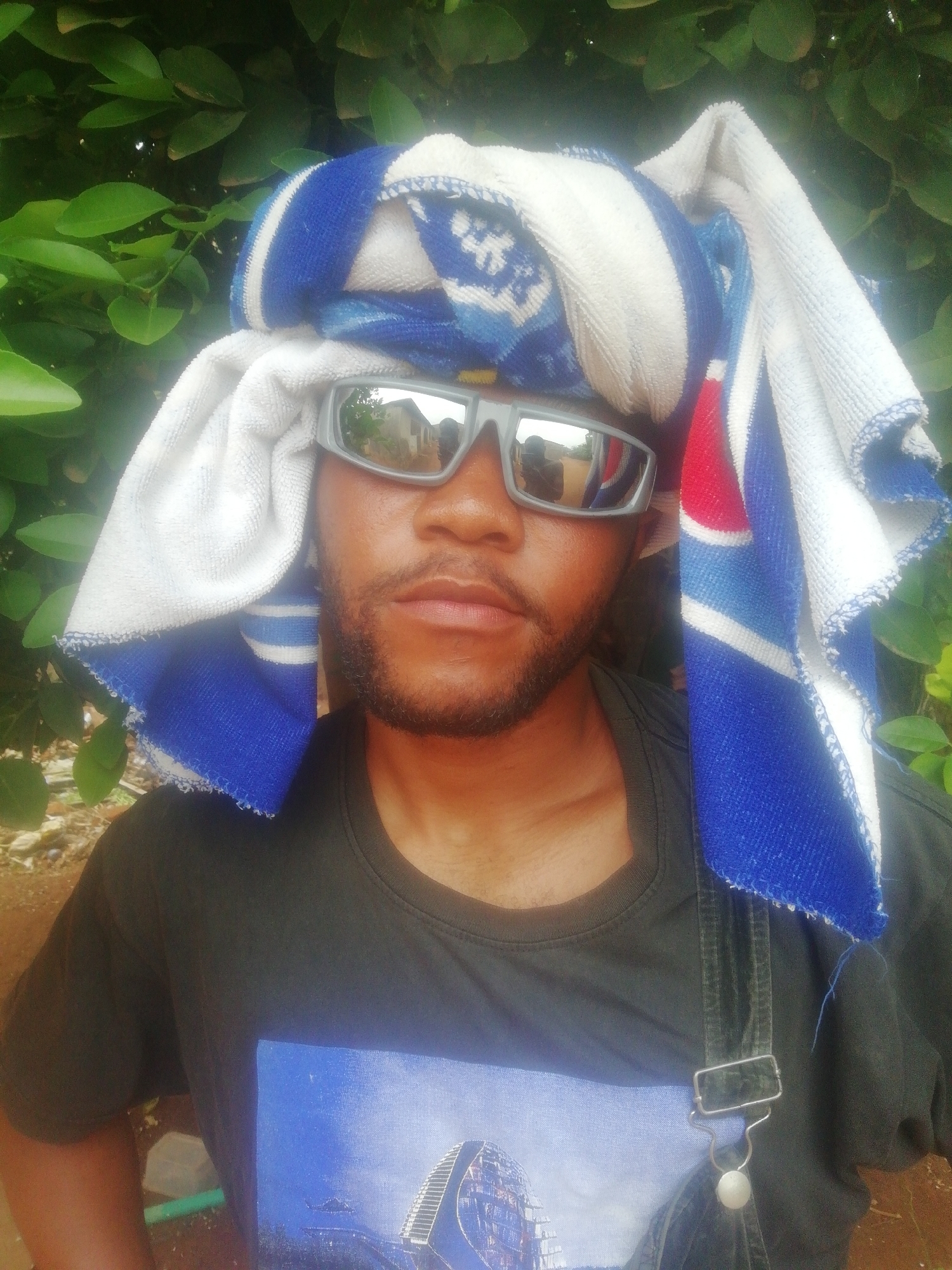 A man is centered in the frame, wearing sunglasses and a towel wrapped around his head. He has a beard and is wearing a dark t-shirt with an image on it. He also appears to be wearing overalls. Green foliage is visible in the background. The image seems to have been taken outdoors in Ikorodu, Nigeria, given the vegetation and possible reflection in the glasses.