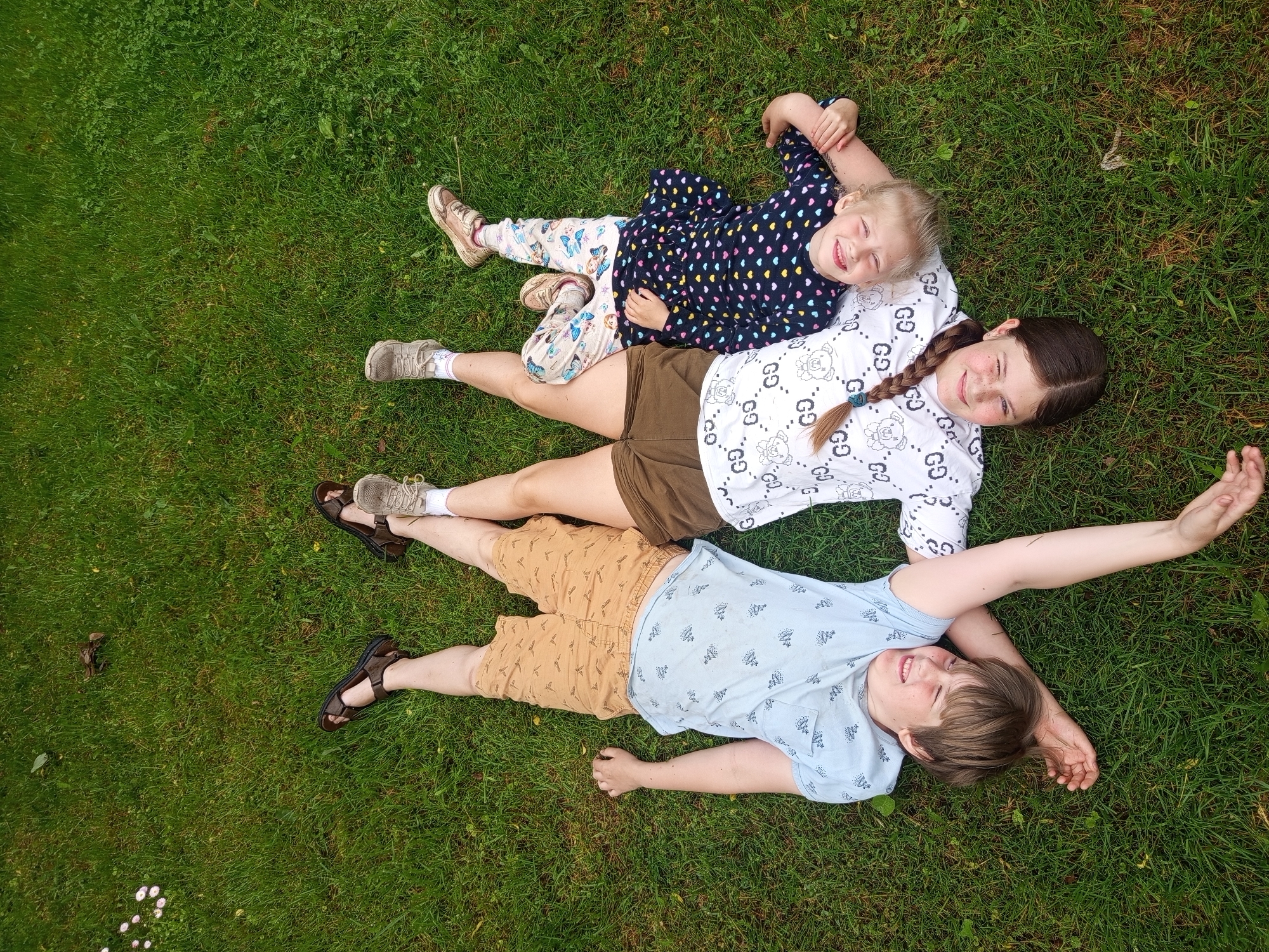 An overhead shot depicts three individuals lying supine on a manicured green lawn, arranged in a pinwheel-like configuration with their heads oriented towards the bottom of the frame, in the city of Unknown, Unknown.
On the left, a male child with light brown hair and a smiling expression, eyes mostly closed, wears a light blue short-sleeved shirt with a faint pattern and tan shorts with a darker repeating motif. His bare feet are visible, and dark brown sandals are positioned nearby in the upper left background.
In the center, a female with long dark brown hair styled in a single braid, secured with a teal elastic, smiles with open eyes. She wears a white short-sleeved t-shirt patterned with repeating "GG" logos and dark olive-green shorts. Light-colored athletic shoes with white socks are on her feet. Her right arm extends towards the male child, her hand resting near his side.
On the right, a younger female child with blonde hair smiles broadly with closed eyes, her head resting on the shoulder of the central female. She wears a dark blue long-sleeved shirt with a colorful repeating heart pattern and light-colored pants featuring a blue butterfly design. Her feet are clad in light-colored athletic shoes.
The ground consists of green grass with interspersed clover and small white and pink blossoms, particularly visible in the upper left quadrant. No other structures or objects are discernible in the immediate vicinity beyond the individuals and the grass.
