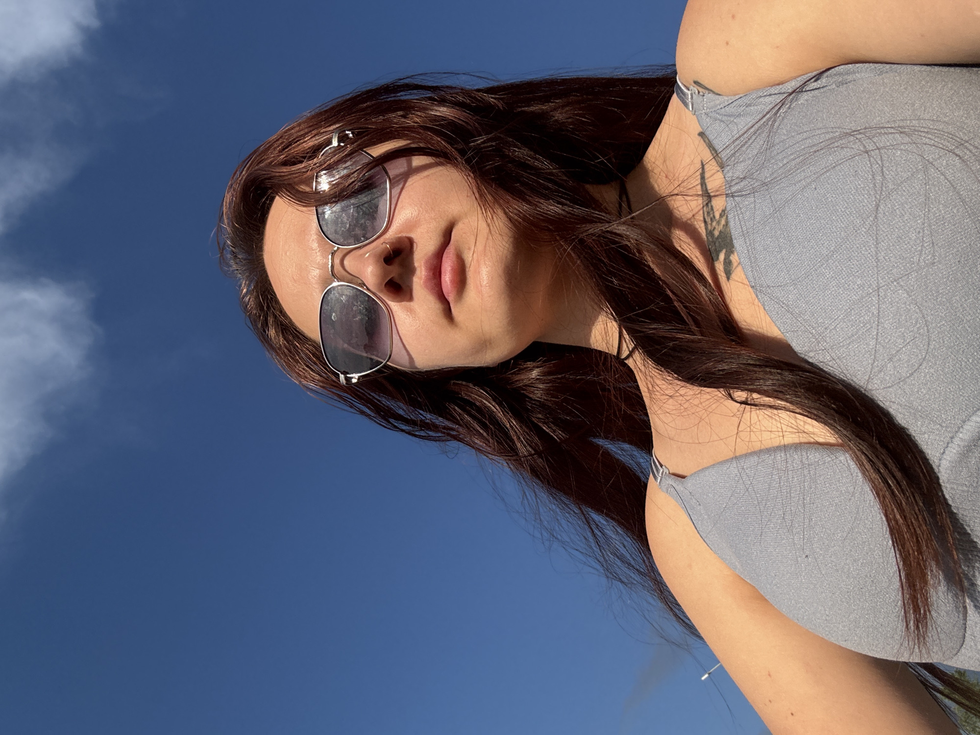 An outdoor, low-angle, medium shot captures a young person from the chest up, looking up at the sky. The person has long, dark brown hair, bangs, and is wearing sunglasses with silver frames and purple lenses. A dark choker necklace is visible around their neck, and they are wearing a grey top with thin straps. A tattoo of a bird is visible on their upper back and shoulder. The background is a clear, bright blue sky with a few wispy white clouds. The image does not provide any specific information to indicate it was taken in Naberezhnye Chelny, Russia.
