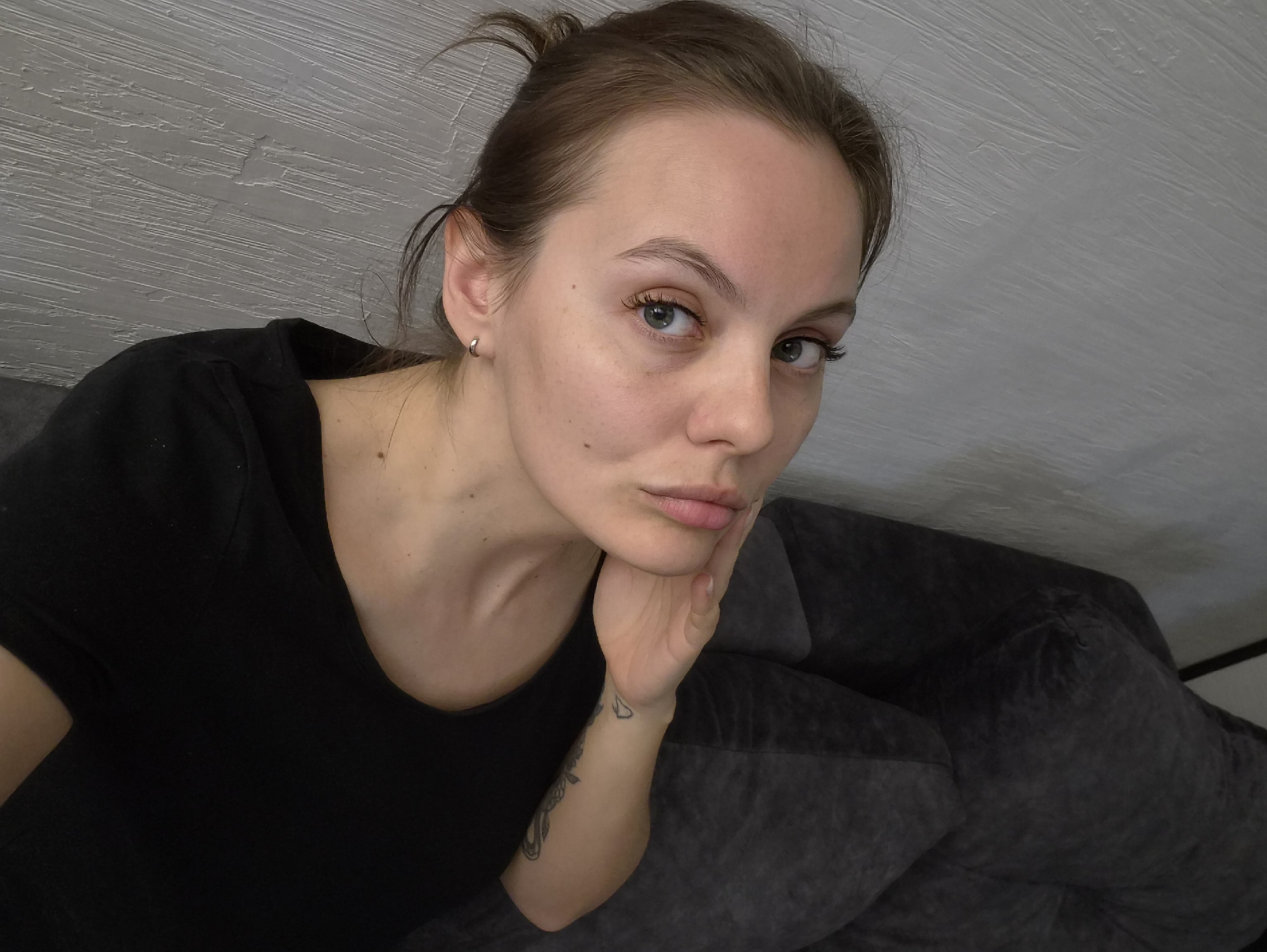 A young adult woman is featured in an indoor selfie, looking directly at the camera with a neutral to slightly pensive expression. Her hair is pulled back in a bun, and she wears a black t-shirt and a small hoop earring in her left ear. Her right hand is resting on her cheek, and a tattoo is visible on her left forearm. The background consists of a textured light grey wall and a dark grey sofa or couch, suggesting a contemporary interior setting. No specific activity beyond taking the photo is apparent. The location context for this image is Chaikovsky, Russia. There is no visible text in the image.