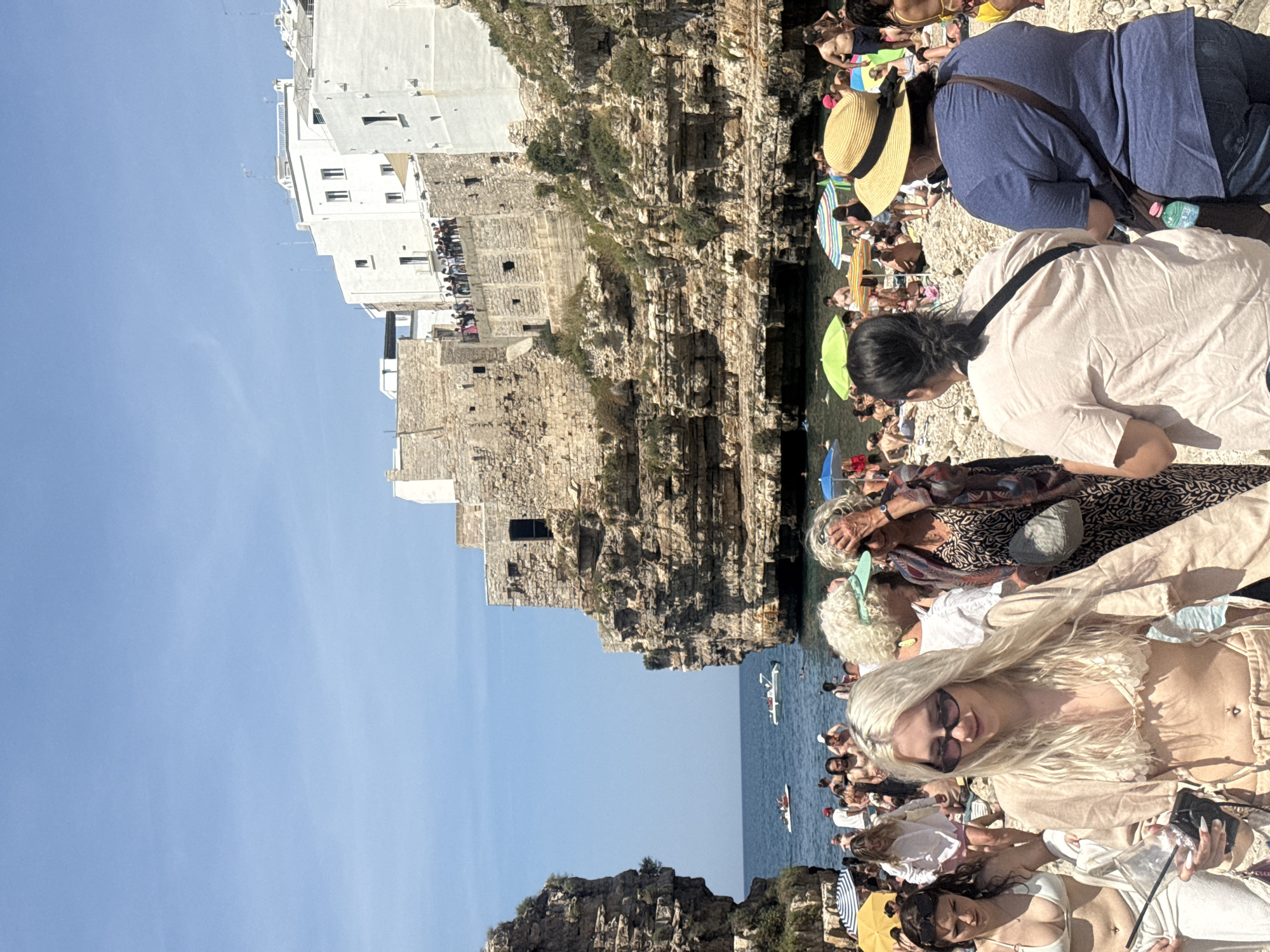 The image depicts a crowded beach scene in Polignano a Mare, Italy. White buildings and stone cliffs form the backdrop to the beach. Numerous people are visible, some sunbathing, others in the water, and some walking along the cliffs. Boats are seen on the water. The sky is clear and blue.