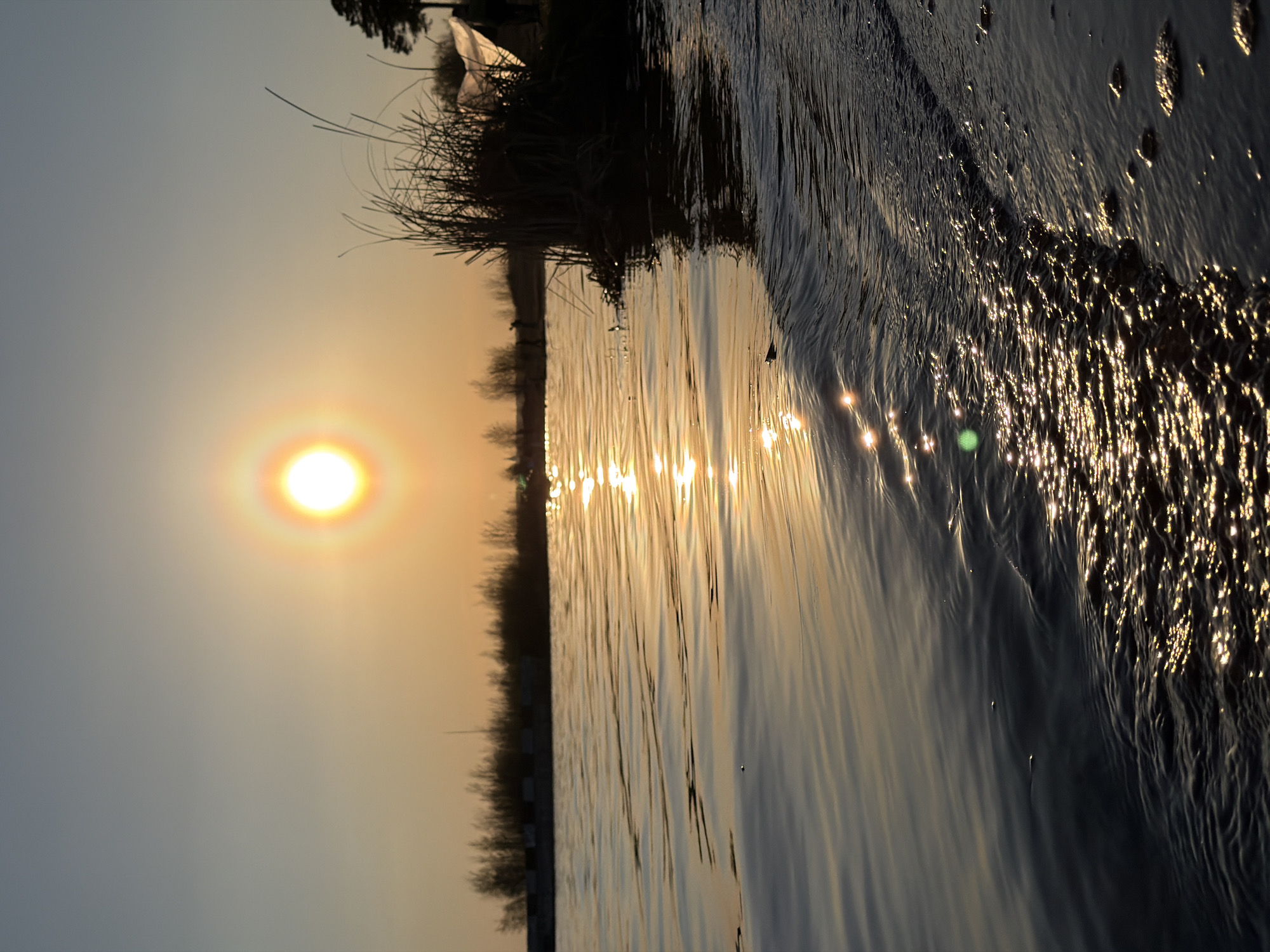 The image depicts a landscape scene, likely taken in Izhevsk, Russia, at sunset. The sun is prominently positioned in the upper left, casting a warm, golden glow across the sky and reflecting brightly on the water's surface. The water takes up a significant portion of the image, with light reflecting off the surface and creating a shimmering effect. Along the horizon, there is a dark silhouette of land, likely a shoreline or a wooded area. There are no people visible in the image. The environment is natural, showcasing a body of water and its surrounding landscape at sunset. The image captures the interaction between light and water.