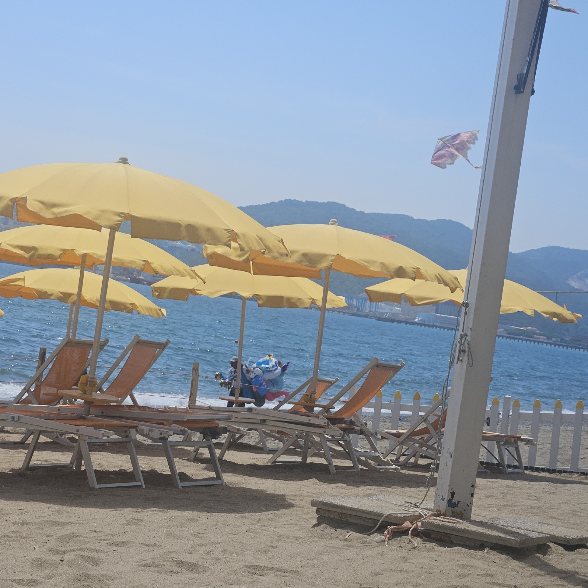 The image depicts a sunny beach scene on a bright day, likely in the city of Savona, Italy, given the presence of numerous yellow umbrellas and lounge chairs lined up on the sand. In the background, the turquoise sea stretches to the horizon, meeting a hazy blue sky. Mountains can be seen in the distance, suggesting a coastal location. A white pole, likely part of a beach structure, is prominently positioned in the foreground. A colorful kite, possibly a child's toy, is visible flying in the air, tethered to the pole or nearby. Several empty lounge chairs are arranged under the umbrellas, indicating a quiet moment on the beach. In the mid-ground, a person is visible near the water's edge, holding a bundle of colorful balloons, possibly a vendor. The overall atmosphere is one of a relaxed summer day at the seaside.