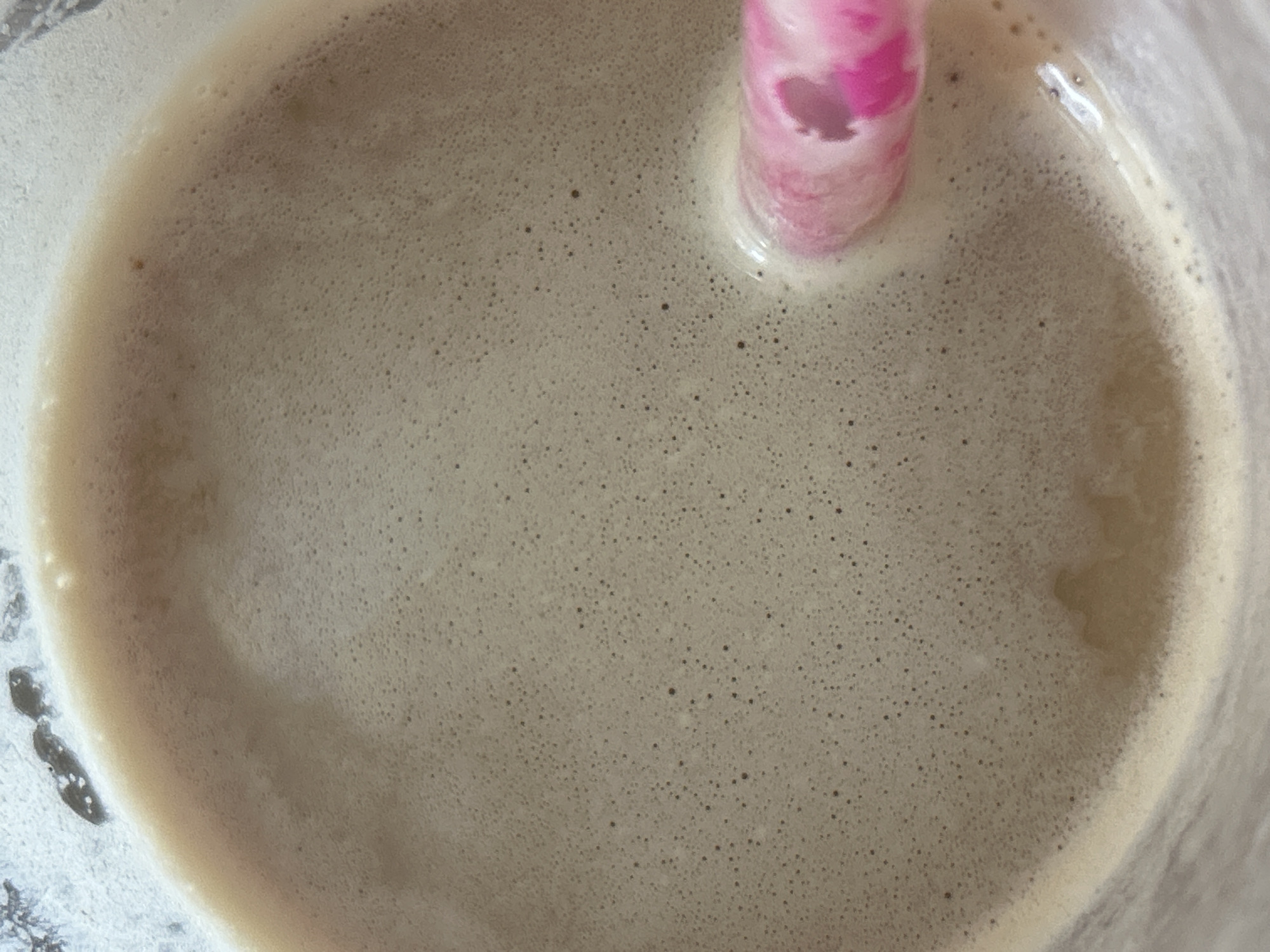 A close-up, overhead shot captures a frothy beverage in a clear glass, with a bright pink straw submerged in the liquid. The drink appears to be a light brown color, possibly coffee or a milkshake, with a fine layer of bubbles on the surface. The lighting is soft, creating a slightly diffused effect. The image does not contain any discernible elements that would suggest a specific location in Plovdiv, Bulgaria.