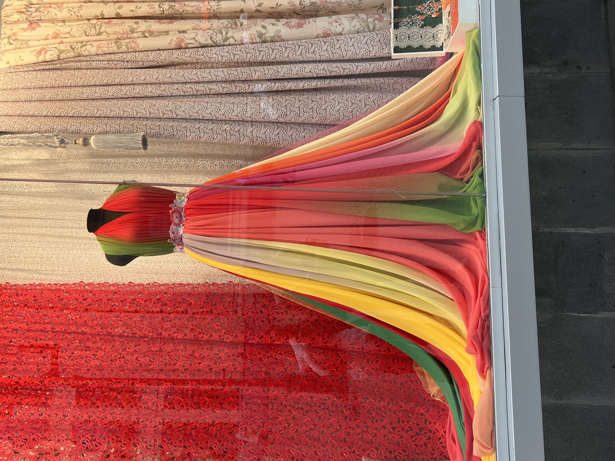 A brightly colored, flowing dress is displayed in a shop window in Yekaterinburg, Russia. The dress, worn by a mannequin, features a halter neckline and a full, floor-length skirt made of sheer fabric in a rainbow of colors, including red, orange, yellow, green, and pink. A jeweled belt cinches the waist of the dress. The dress is displayed against a backdrop of patterned curtains and red lace fabric. To the right of the dress, on a shelf, is a small white box containing jewelry and other decorative items. The shop window is made of glass, and the exterior wall of the building, constructed of dark stone blocks, is visible below the window.