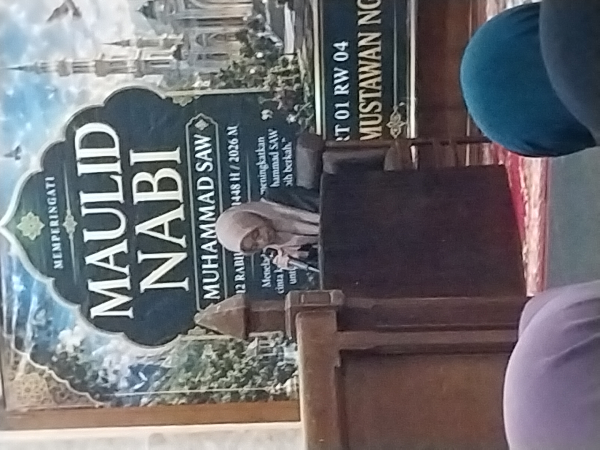 The image depicts an indoor event, likely a religious gathering, taking place in Nglegok, Indonesia. In the foreground, a woman wearing a light brown hijab is speaking into a microphone, positioned behind a wooden podium. She is positioned to the left of a dark-colored rectangular structure, possibly a lectern or display. Behind her, a large banner features prominent text that reads "MAULID NABI MUHAMMAD SAW" and includes dates "12 RABIUL AWAL 1448 H / 2026 M". The banner also displays an illustration of a mosque with minarets and birds in flight. To the right of the banner, another sign reads "RT 01 RW 04 MUSTAWAN NG". In the lower portion of the image, the heads of two individuals wearing head coverings are visible, one in light purple and another in dark blue, suggesting they are seated attendees. The overall setting appears to be a community hall or mosque, with red and white patterned carpeting visible on the floor.