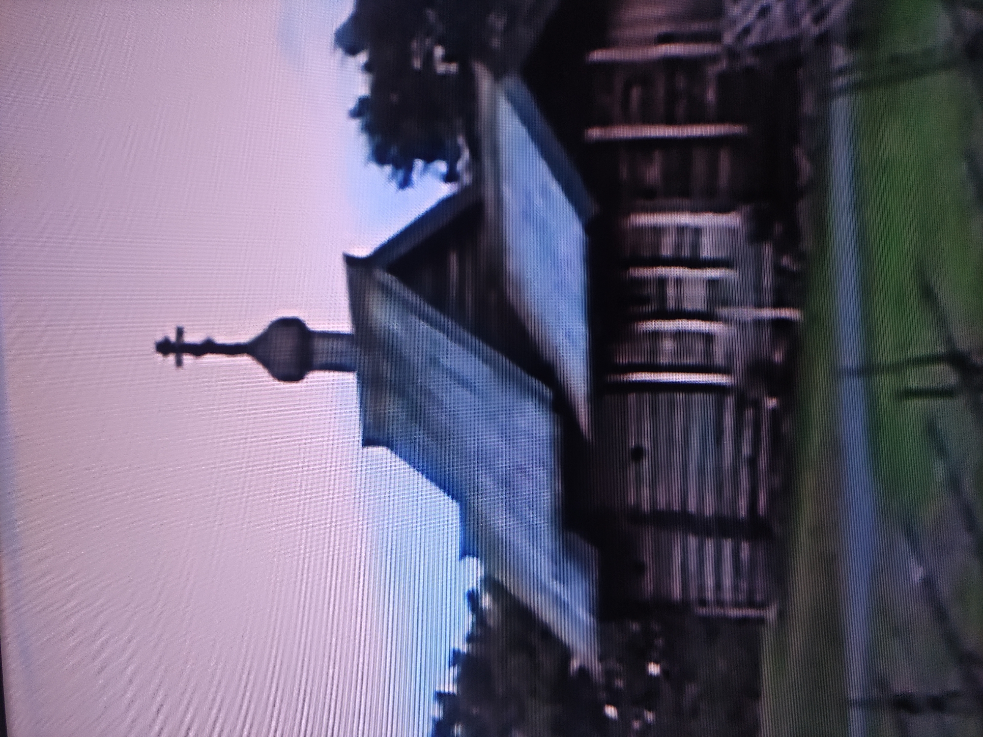 The image depicts a wooden structure with a distinctive architectural style, likely an Orthodox church or chapel, situated in Pavlodar, Kazakhstan. The building has a steeply pitched roof covered with wood and is surmounted by a small dome topped with a cross. The walls are constructed of horizontal wooden planks or logs. Adjacent to the structure is an area with green grass and a fence or other boundary marker. The sky is light in color. The photo has some visual noise or artifacts, possibly from the recording or scanning of an older image. The orientation of the image is sideways.