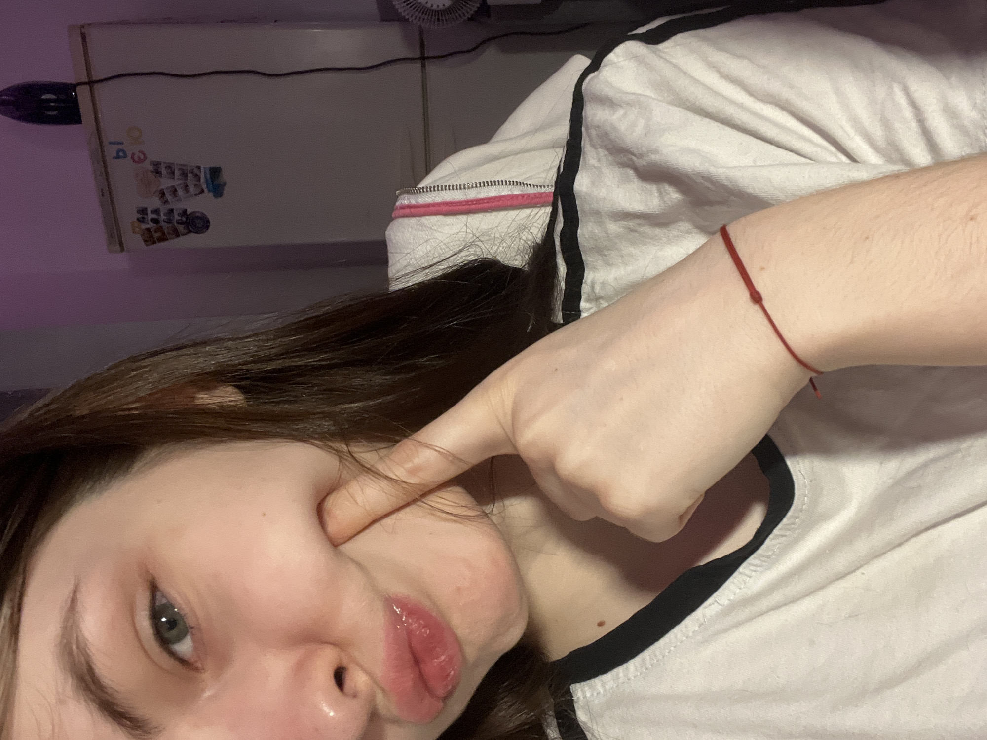 A selfie of a young woman with brown hair, light skin, and grey-green eyes. She is pressing her finger into her cheek, distorting her face. She has full lips with a slight sheen. She is wearing a white t-shirt with black trim around the neckline. A thin red bracelet is visible on her left wrist. The background includes a white wall with stickers or magnets, a fan, and a power cord. The lighting is somewhat dim, with a purple hue on the left side of the image. There is no indication that this image was taken in Saint Petersburg, Russia.