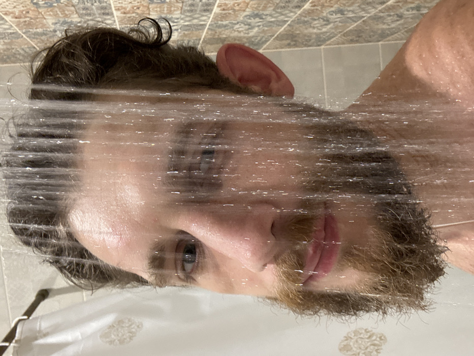 The image presents a close-up, rotated portrait of a person taking a shower. The image is rotated 90 degrees clockwise, so the person's head is tilted to the right, and what would normally be vertical elements (like falling water) appear horizontal across the frame.
The central figure is a person, likely male, with dark brown, curly hair visible on top of their head and a prominent beard and mustache that appear to be a mix of brown and auburn tones. Streams of water from the shower are actively falling across the person's face, creating numerous shimmering, horizontal lines that partially obscure facial features but add a sense of motion and wetness.
Despite the water, the person's eyes are open and gaze directly towards the viewer, appearing light in color (possibly blue or grey). Their expression is neutral. The skin on their face, including the forehead, cheeks, nose, and lips, appears wet and slightly glistening. A portion of their ear is visible at the top of the head.
In the background, light-colored bathroom tiles with a subtle, decorative pattern are visible, positioned at the top and to the right of the person's head due to the image's rotation. Below the person's chin and chest, a white shower curtain with a faint, almost floral-like pattern can be seen. The overall scene captures a candid moment during a shower, emphasizing the water and the person's direct gaze. No text is visible in the image.