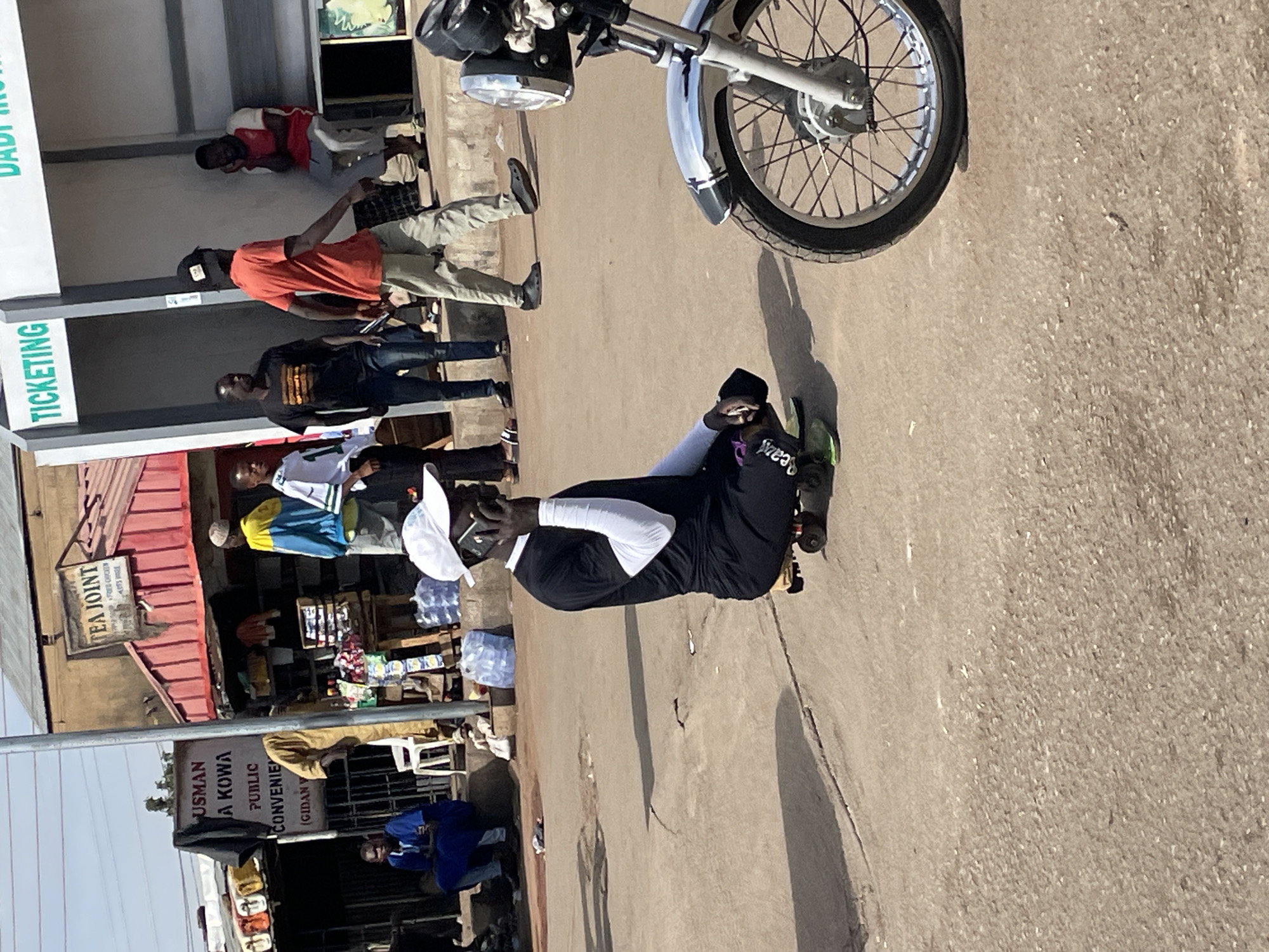 The image depicts a street scene in Jos, Nigeria, featuring a variety of elements. A motorcycle's front wheel and headlight dominate the upper right. Several people are present: one in a black top and white sleeves talking on a cell phone, another in an orange shirt and tan pants, and others in various attire. Some stand near a building marked "TICKETING" and "DADI." A "TEA JOINT" sign is visible on another building, along with a sign for "USMAN A KOWA PUBLIC CONVENIENCE." Additional buildings and vendor displays line the street. The scene is bathed in sunlight, casting distinct shadows.