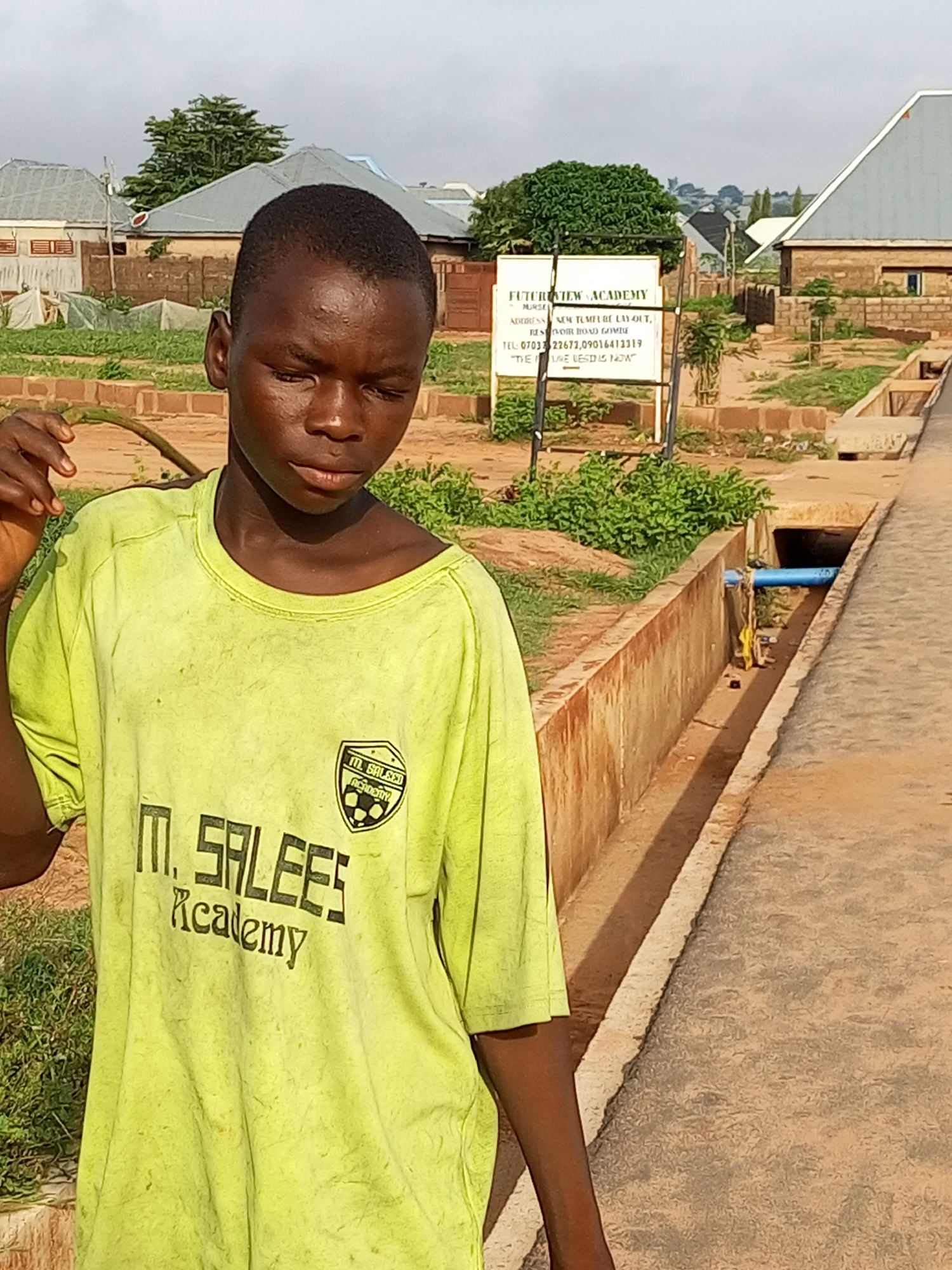 A young boy with dark skin and short black hair stands in the foreground, facing slightly to his left with his eyes closed and head tilted down. He is wearing a faded, bright yellow t-shirt with black text and a logo on the left chest. The text on the shirt reads "M. SALEES Academy". In the background, a white sign with black text is visible. The sign appears to be a school sign, with "FUTUREVIEW ACADEMY" at the top and "NURSE" below it. Further details on the sign include an address and phone number, and the slogan "THE FUTURE IS NOW". Behind the sign, there are buildings with gray roofs and light-colored walls, surrounded by green vegetation. To the right of the boy, there is a concrete drainage ditch with a blue pipe running across it, and a paved walkway. The overall environment appears to be an outdoor school or community area in Tempure, Nigeria.