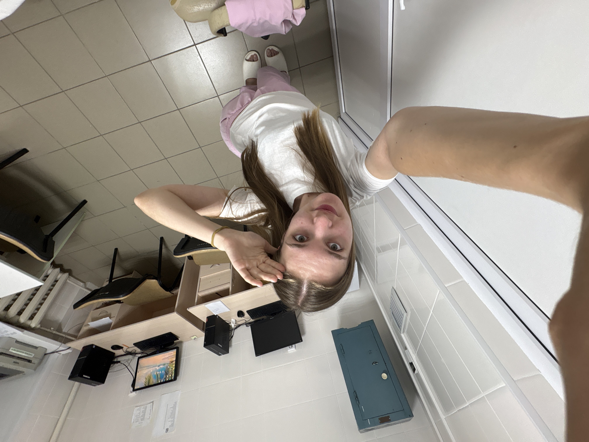 A person with long brown hair, wearing a white t-shirt and pink pants with white slippers, is lying on the floor, taking a selfie. The location appears to be a room with white tiled walls and a tiled floor. A desk with a computer monitor, keyboard, mouse, and CPU is visible. There are chairs, a blue metal cabinet, and a printer in the room. The city is Perm, Russia.