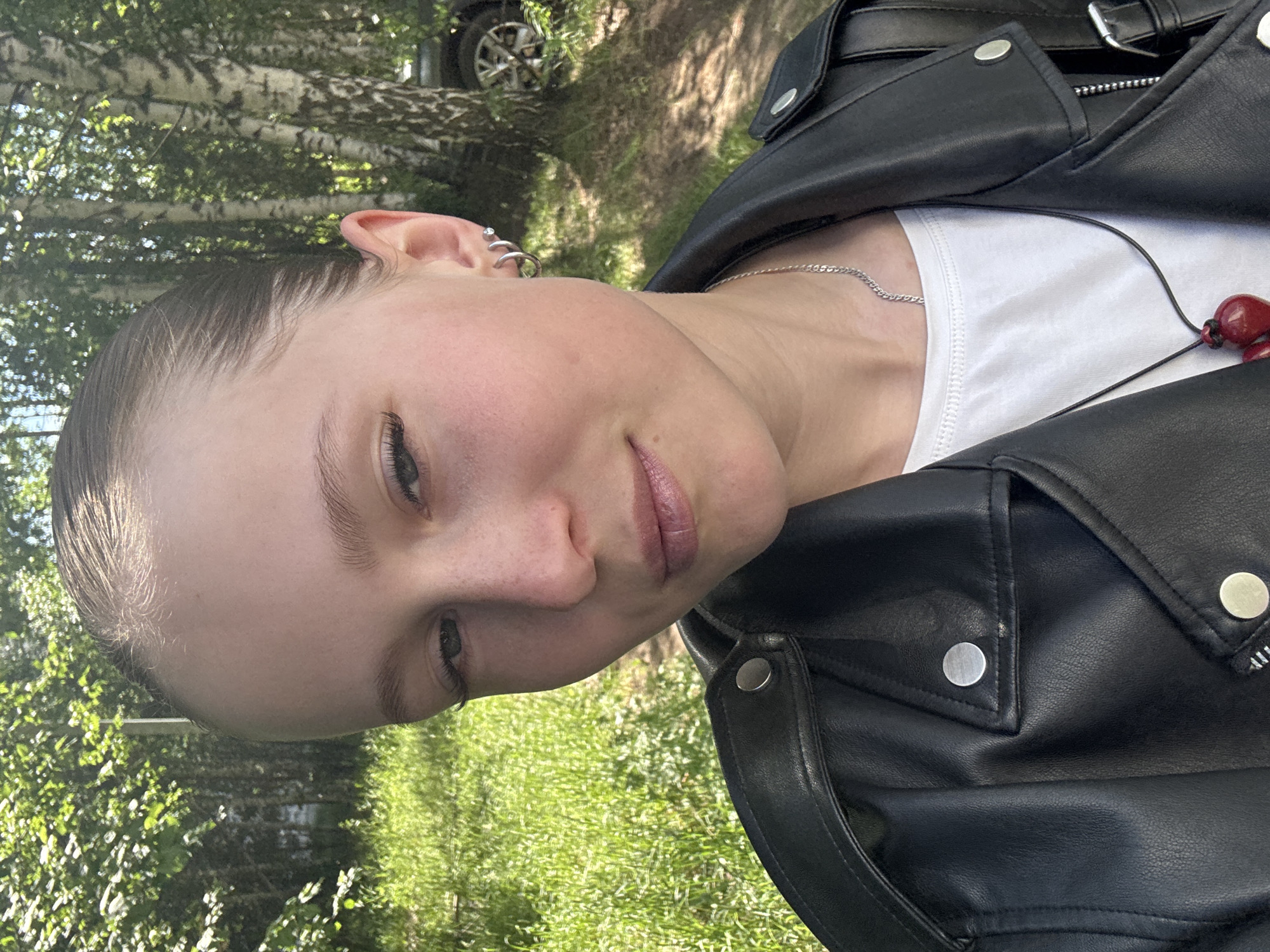 The image is a close-up, outdoor portrait of a young female, appearing to be a selfie. She has fair skin, light brown hair slicked back tightly, prominent eyebrows, and hazel or greenish-grey eyes looking directly at the camera. Her expression is neutral with slightly parted, naturally colored lips. Her left ear is visible, adorned with two small silver hoop earrings and one small silver stud piercing.
She is wearing a black biker-style faux leather jacket with large lapels and silver snap buttons visible on the shoulder epaulet and a small chest pocket flap. Beneath the jacket, a plain white crew-neck t-shirt is visible. Around her neck, she wears a thin silver chain necklace and a longer black cord necklace with a glossy red, heart-shaped pendant.
The background is an outdoor, sunlit natural environment. Several mature birch trees with characteristic white bark and dark markings are prominently featured, suggesting a forest edge or park. Lush green grass covers the ground. In the far right background, partially obscured by the trees, a dark-colored vehicle (likely an SUV or car) is visible, indicating proximity to a road or parking area. The lighting is bright natural sunlight.
The image itself does not contain any visual cues or specific landmarks to forensically confirm the city of Chaikovsky, Russia.