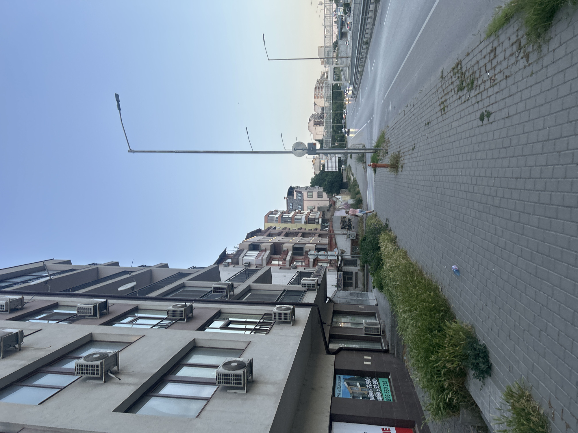 The image captures a street scene in Varna, Bulgaria, likely in the evening, judging by the lighting and the clear blue sky. On the left, a multi-story apartment building with numerous air conditioning units visible on its exterior dominates the foreground. The building has several floors with windows and balconies. A sidewalk made of gray interlocking pavers runs along the base of the building, bordered by a strip of overgrown grass and weeds. A plastic bottle lies on the sidewalk.
The sidewalk leads to a street or highway, with a pedestrian crossing and a fire hydrant visible near the intersection of the sidewalk and the road. A solitary figure, a person in a pink shirt and blue shorts, walks on the sidewalk away from the camera, towards the street.
The road is a multi-lane thoroughfare with cars visible in the distance, some with their headlights on. Streetlights line the road, with one prominent lamp post in the middle ground of the image. In the background, other buildings and structures, including a pedestrian bridge, are visible. The overall impression is that of a modern urban environment.