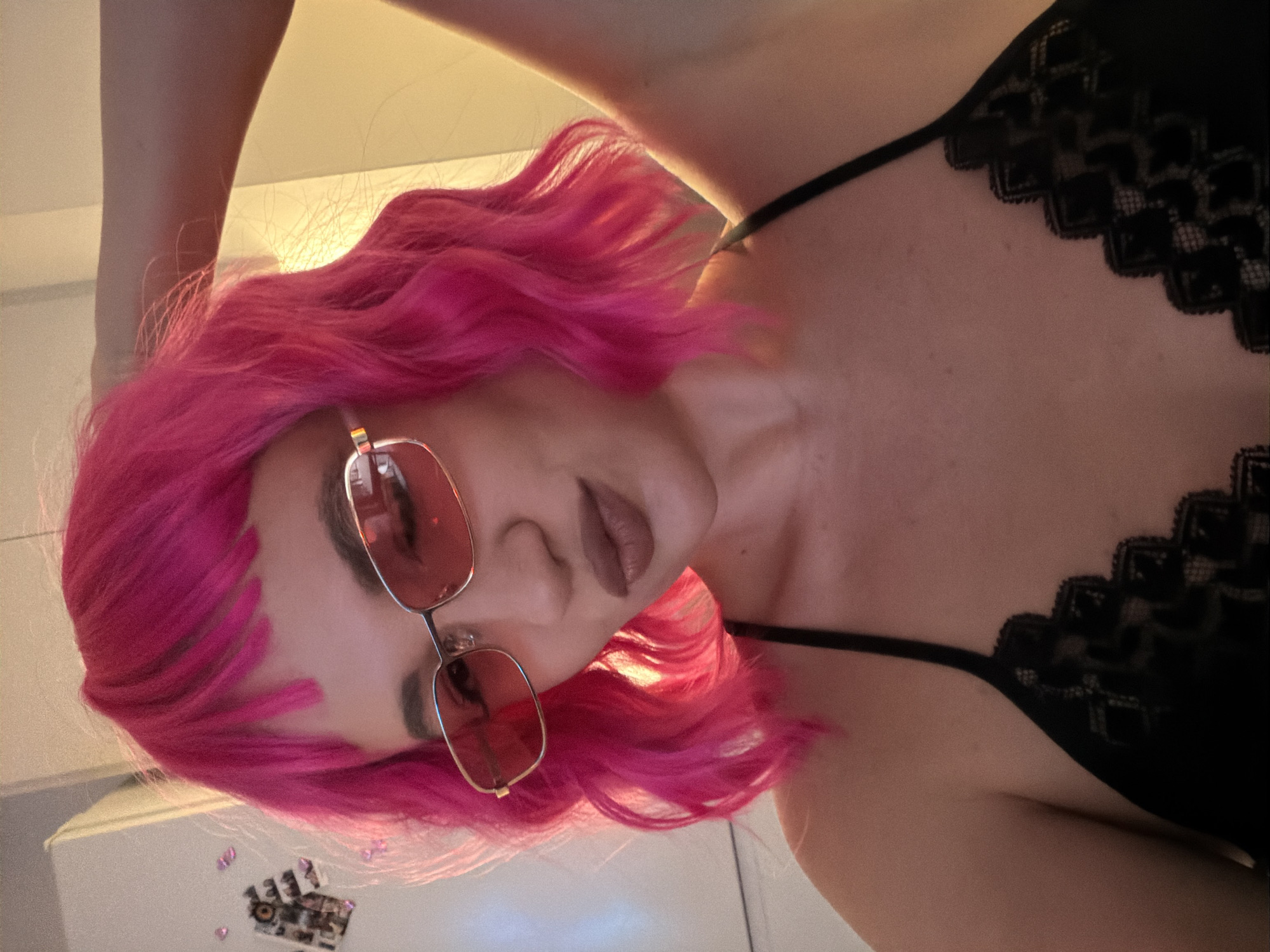 A woman with bright pink wavy hair and light skin is posing for a picture. She is wearing pink-tinted sunglasses with gold frames and a black lace-trimmed top with thin straps. The background shows a plain white surface. There is a white cabinet with some small, square, possibly decorative items. The specific location in Belgrade, Serbia, cannot be determined from the image alone.