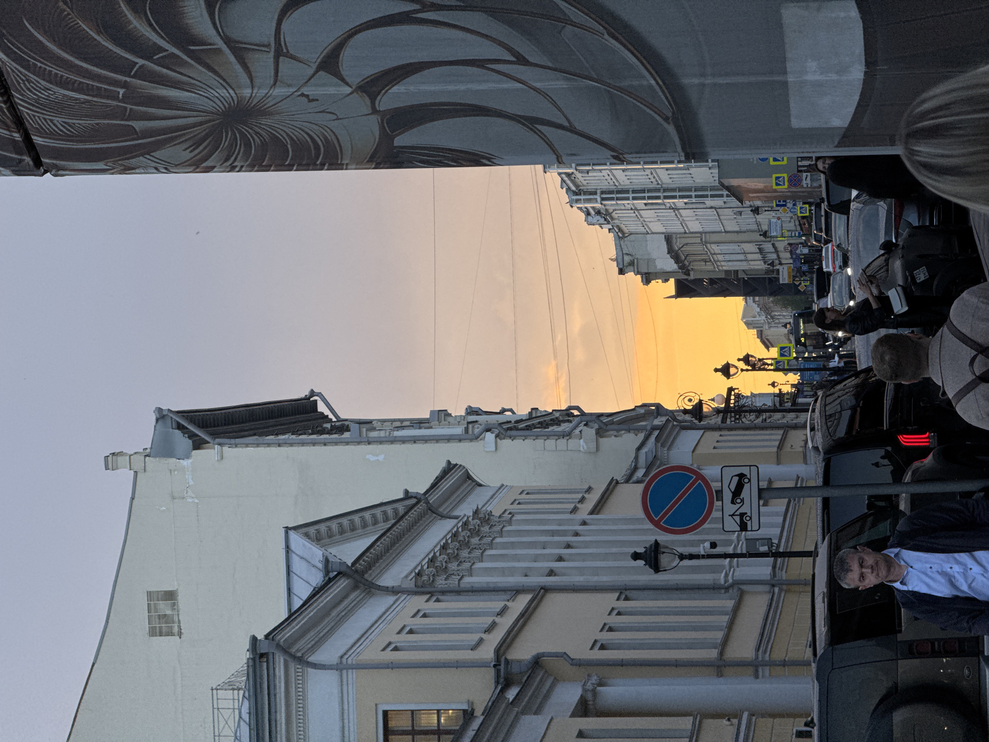 The image shows a street scene in Moscow, Russia, during a sunset. Buildings with ornate facades line both sides of the street, creating a narrow perspective towards a vibrant orange sky. People are present on the street and partially visible, alongside a line of parked cars and street furniture, including street lamps and traffic signs with parking restrictions. The composition appears to be taken from a point of view that includes part of an adjacent structure with a distinctive, swirling artistic pattern.